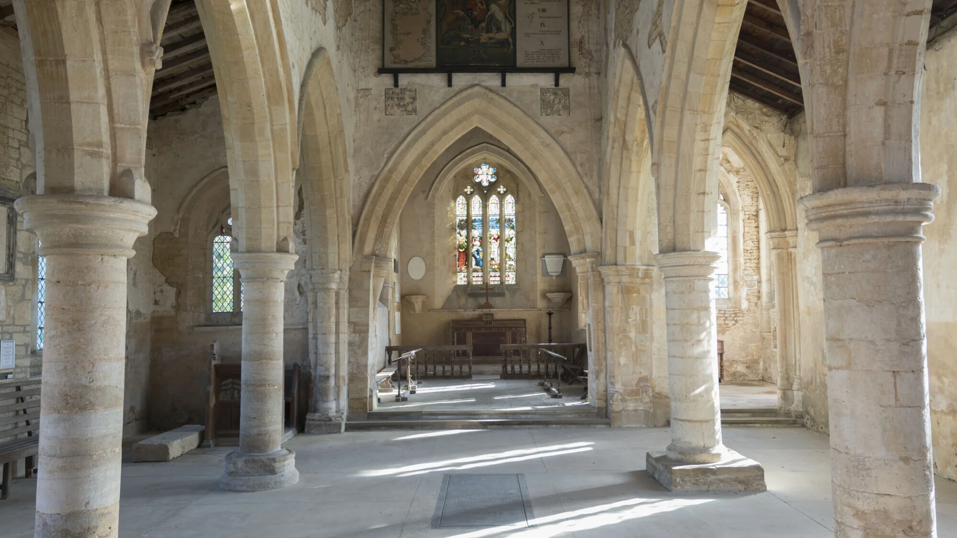 Interior of All Saints, Aldwincle, a perpendicular Gothic church, with some Norman features. The image looks down the nave towards the chancel. The nave is divided into three bays, by this columns with pointed arches. Another pointed arch separates the chancel from the nave. There are no pews or other items of furniture.