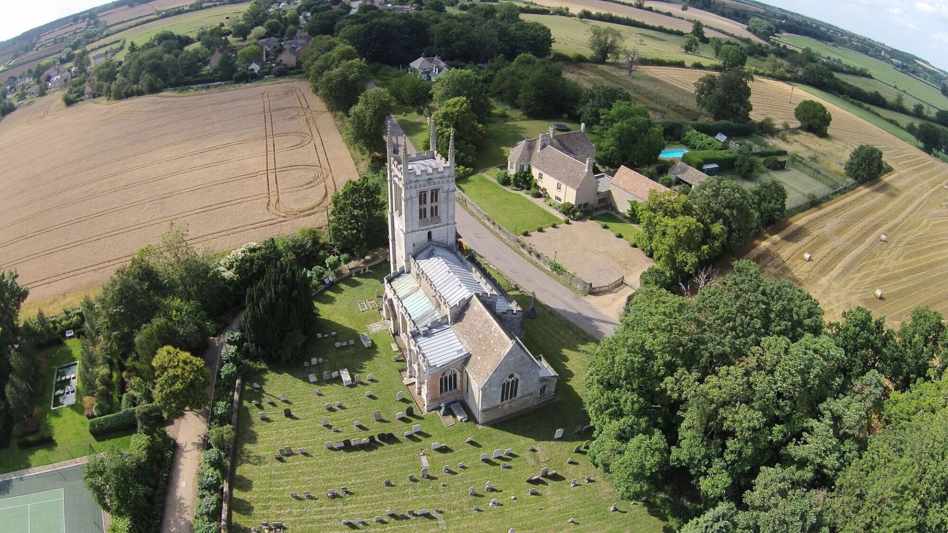 Aerial view of All Saints, Aldwincle, a medieval perpendicular Gothic church, with tall, pinnacled tower, three-bay nave and chancel. This church sits in a green churchyard and is surrounded by brown fields, dark green hedgerows and lush trees.
