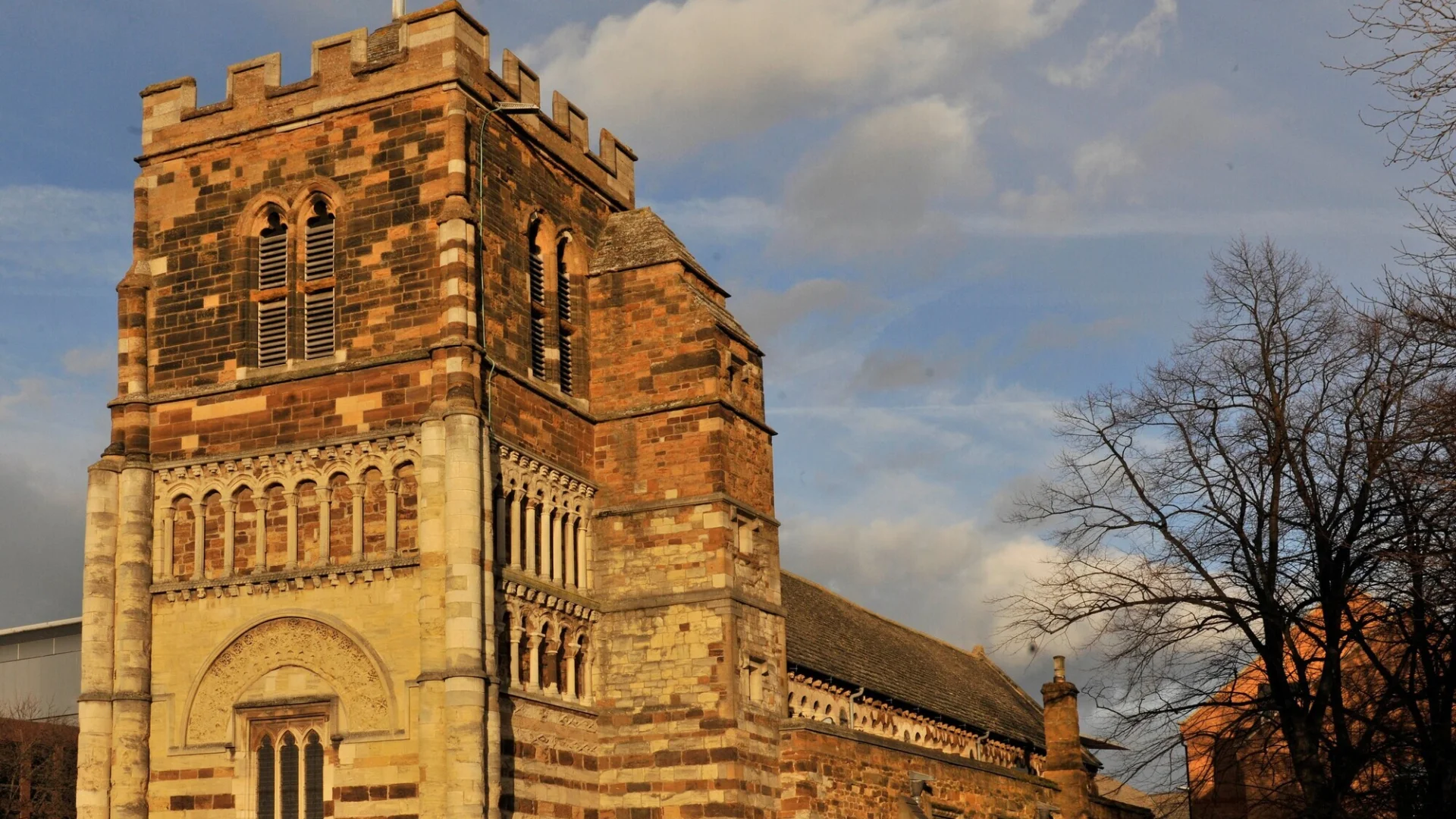 The orange, red and brown ironstone exterior of the squat Norman church of St. Peter's, Northampton, illuminated by the setting sun. The sky behind this imposing building is blue, speckled with clouds and the black silhouettes of bare trees can be seen in the background. The image is dominated by the crennellated tower, on which the stonework looks like a patchwork of materials and architectural periods.