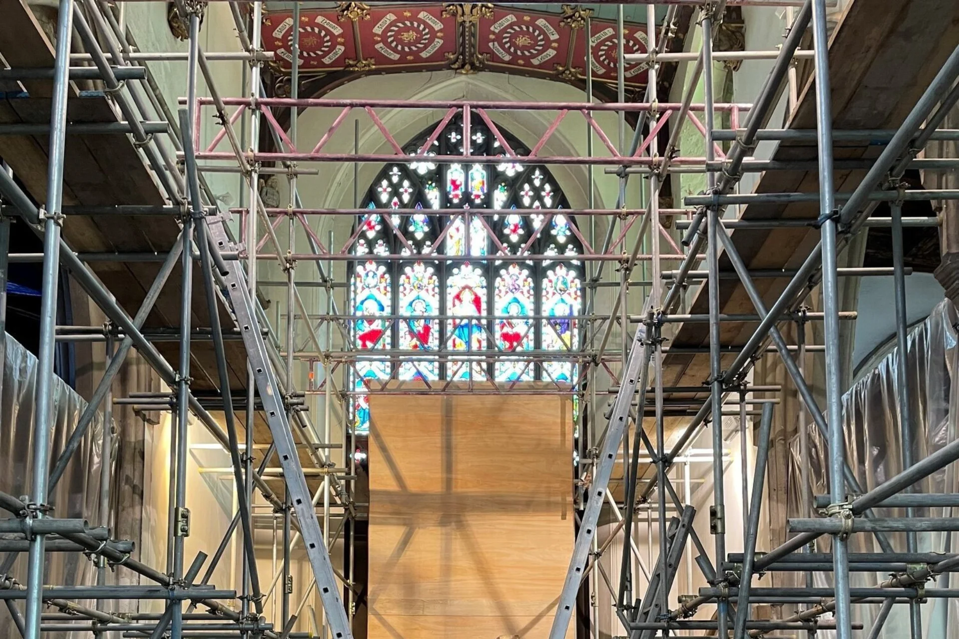 St Peter's Church in Sudbury, looking down the central nave, with floor to ceiling scaffolding and a striking arched stained glass window at the end, partly covered in wood for its protection.