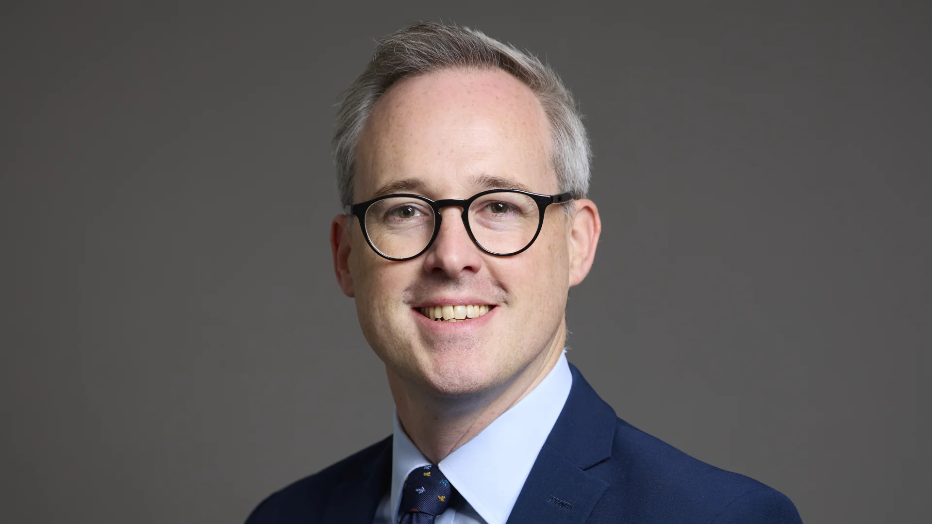 A gentleman wearing glasses poses in front of a plain background. He has a navy blue suit jacket, pale blue shirt and a dark blue patterned tie.