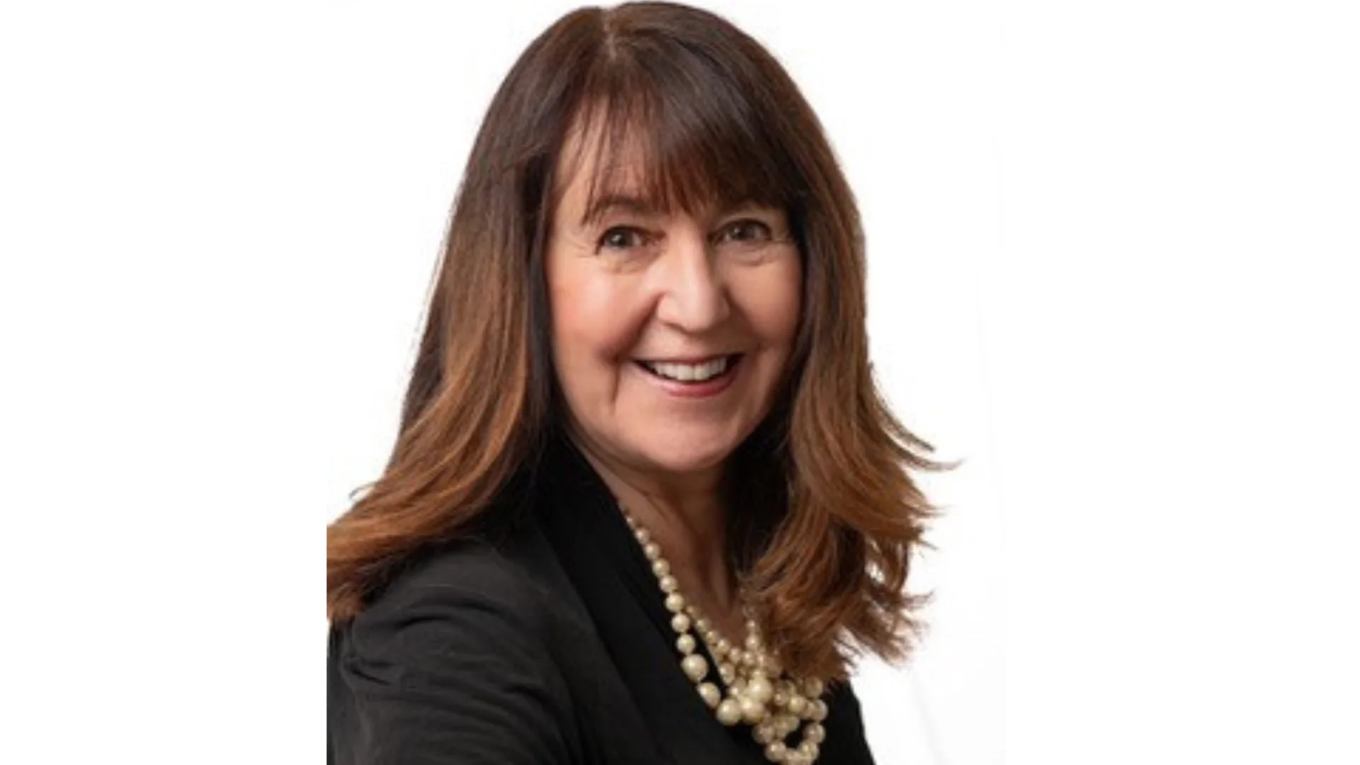 A woman smiles at the camera with a plain white background. She has brown hair, a pearl necklace on and a black suit.