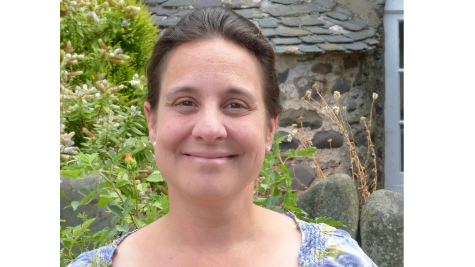 A woman stands in front of a stone building with shrubbery and a stone wall. She has brown hair tied back and wears a lilac floral top.