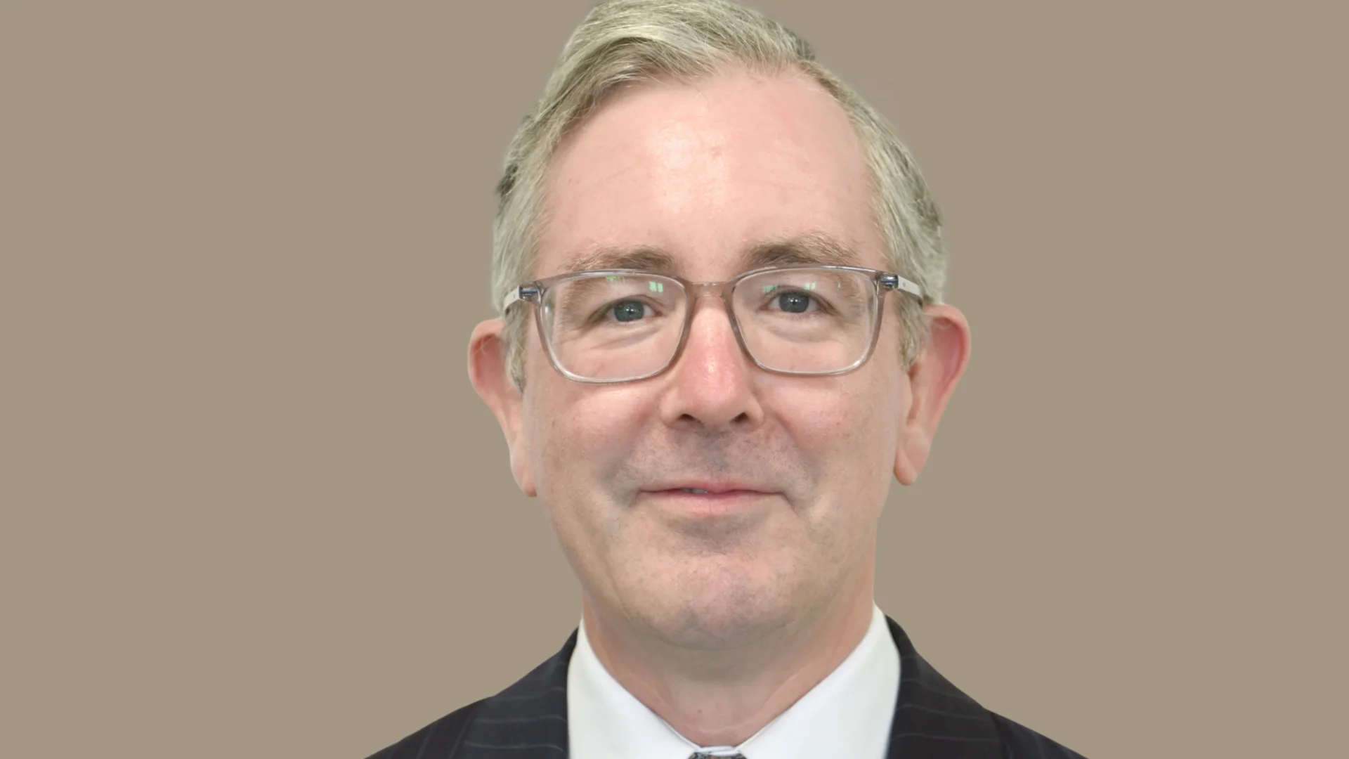 A gentleman stands with a plain background, he is wearing a black suit jacket, patterned tie and white shirt. He also wears glasses.