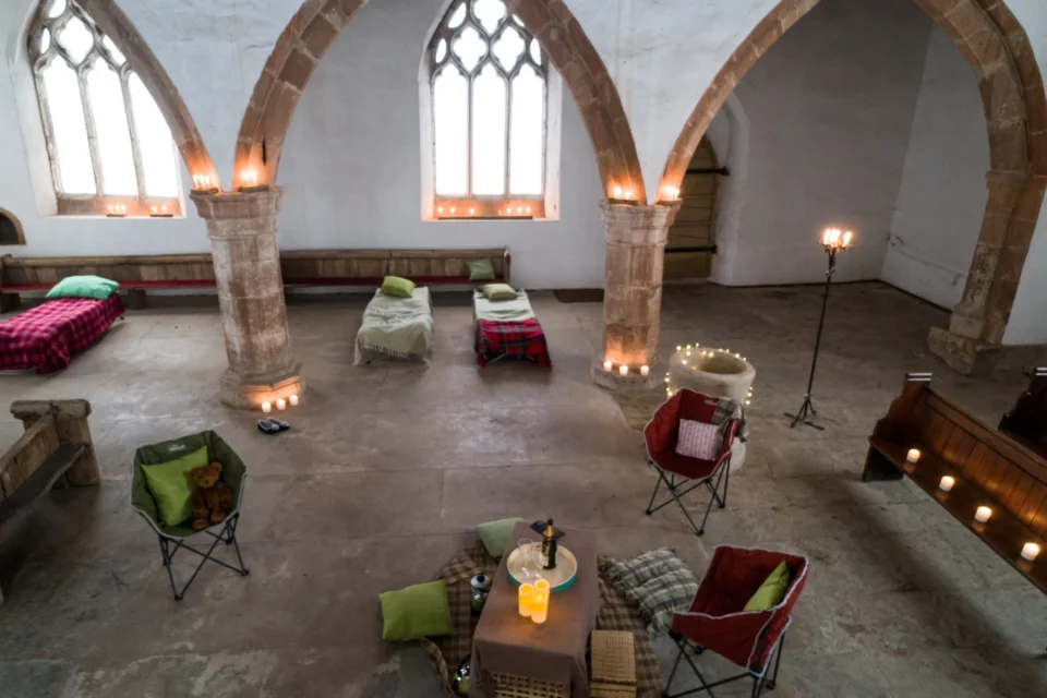 white washed high ceilinged church interior with arches, camp beds and camp chairs with cosy blankets