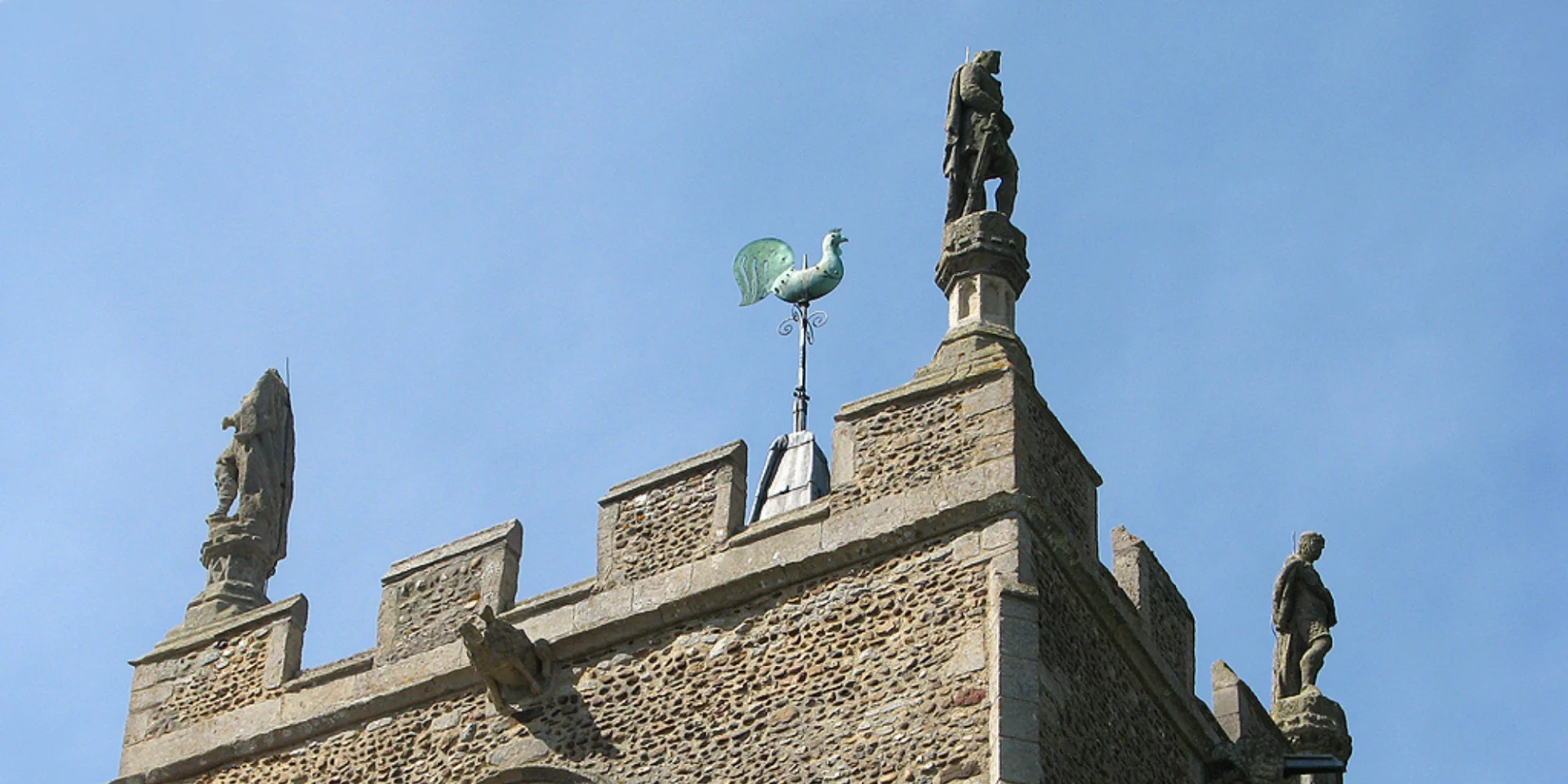 A crenelated church tower with a statue of a medieval king on each corner and a weathervane of a cockerel in the centre.