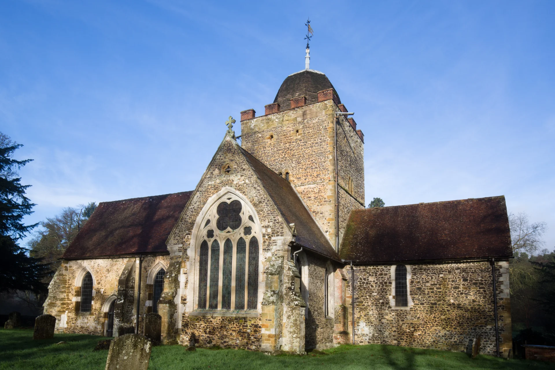 Church exterior with unusual tower with cupola, east window with quatrefoil in front of blue sky in green churchyard.