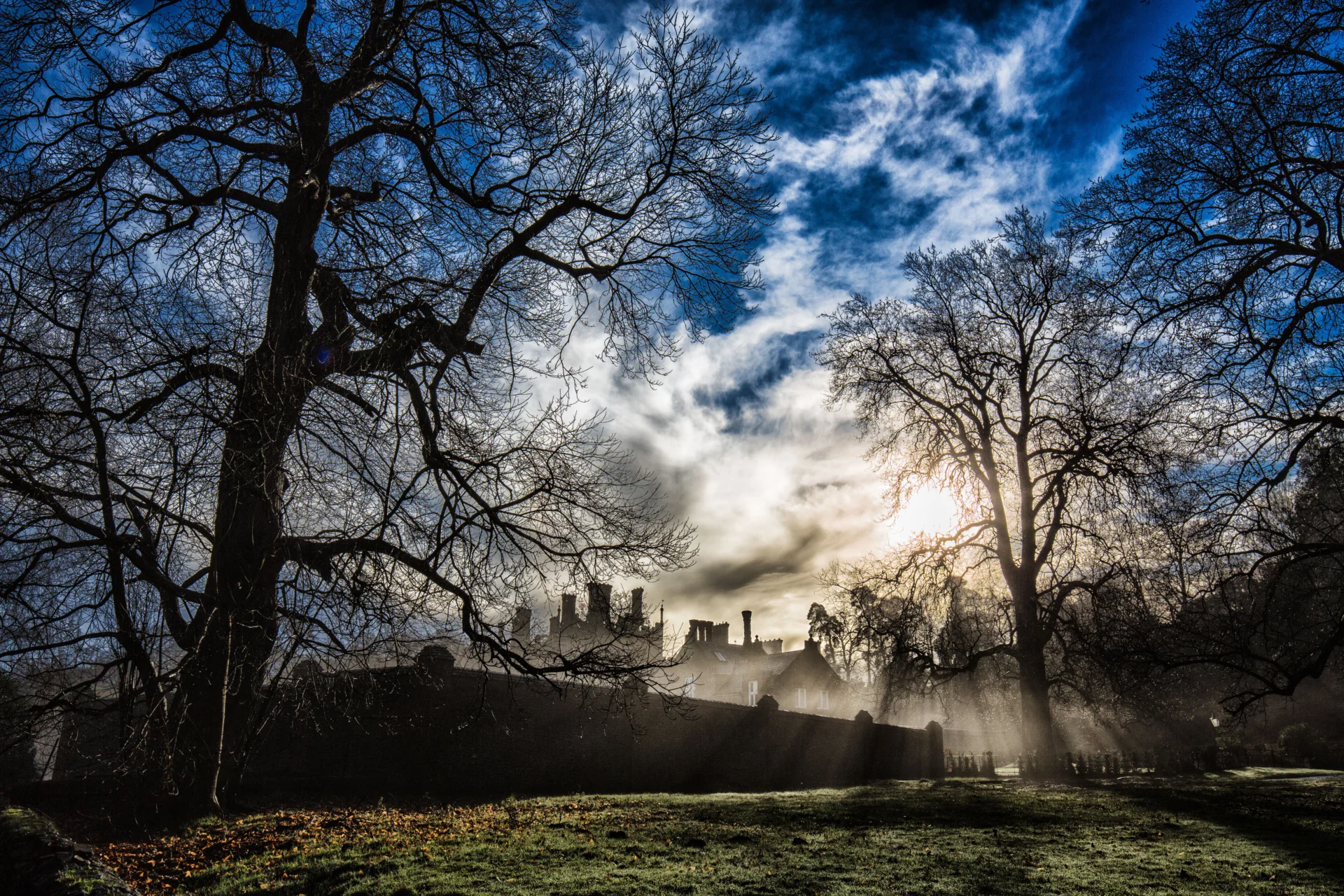 Silhouette of a manor house seen against a dramatic sunrise. Rays of sun part white clouds and blue sky.
