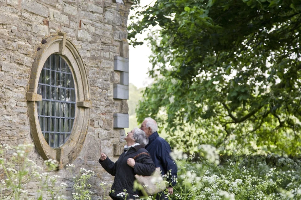 a man and woman explore the outside of a stone church, looking specifically at a round glass porthole window