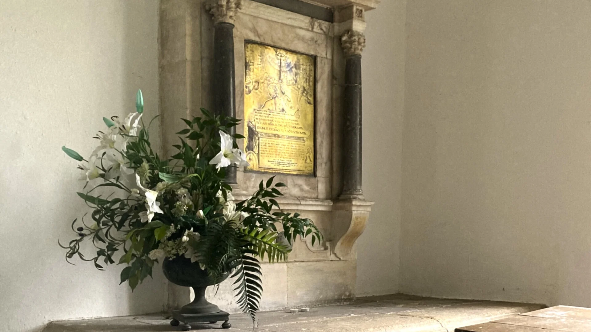 Stone monument tomb-chest in the chancel of Alton Priors with a brass wall-plate above commemorating William Button and portraying him being greeted at the gates of heaven. A vase of artificial lilies sits atop the tomb,