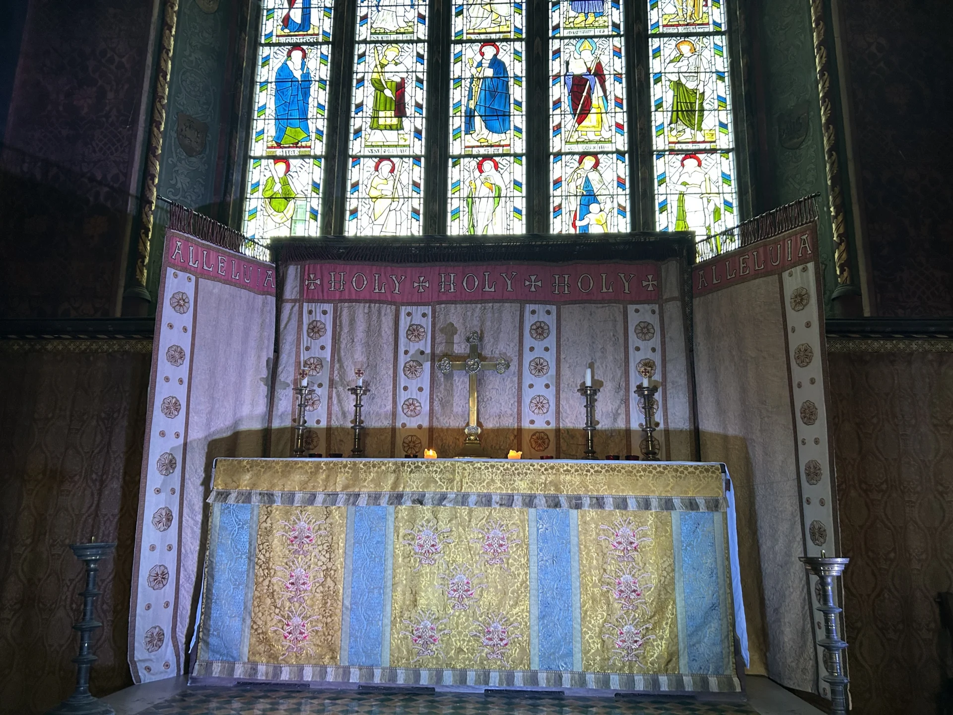 Photograph of altar curtains featuring embroidery of roses and the text "alleluja, holy, holy, holy, alleluja" and altar with cross and candles on, in front of stained glass window depicting saints.