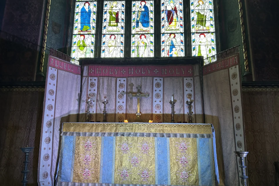Photograph of altar curtains featuring embroidery of roses and the text "alleluja, holy, holy, holy, alleluja" and altar with cross and candles on, in front of stained glass window depicting saints.