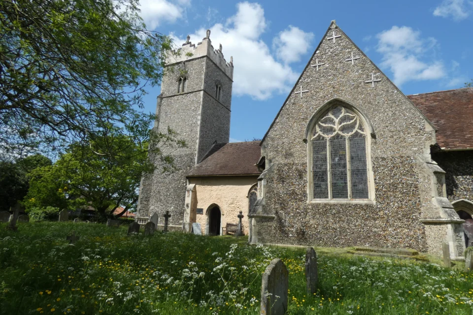 We can see the exterior of St. Peter's, Claydon, a simple flint rubble church with crennelated tower and attractive window tracery sat in a lush green churchyard dotted with a few headstones. The image is taken on a sunny day with bright blue skies.