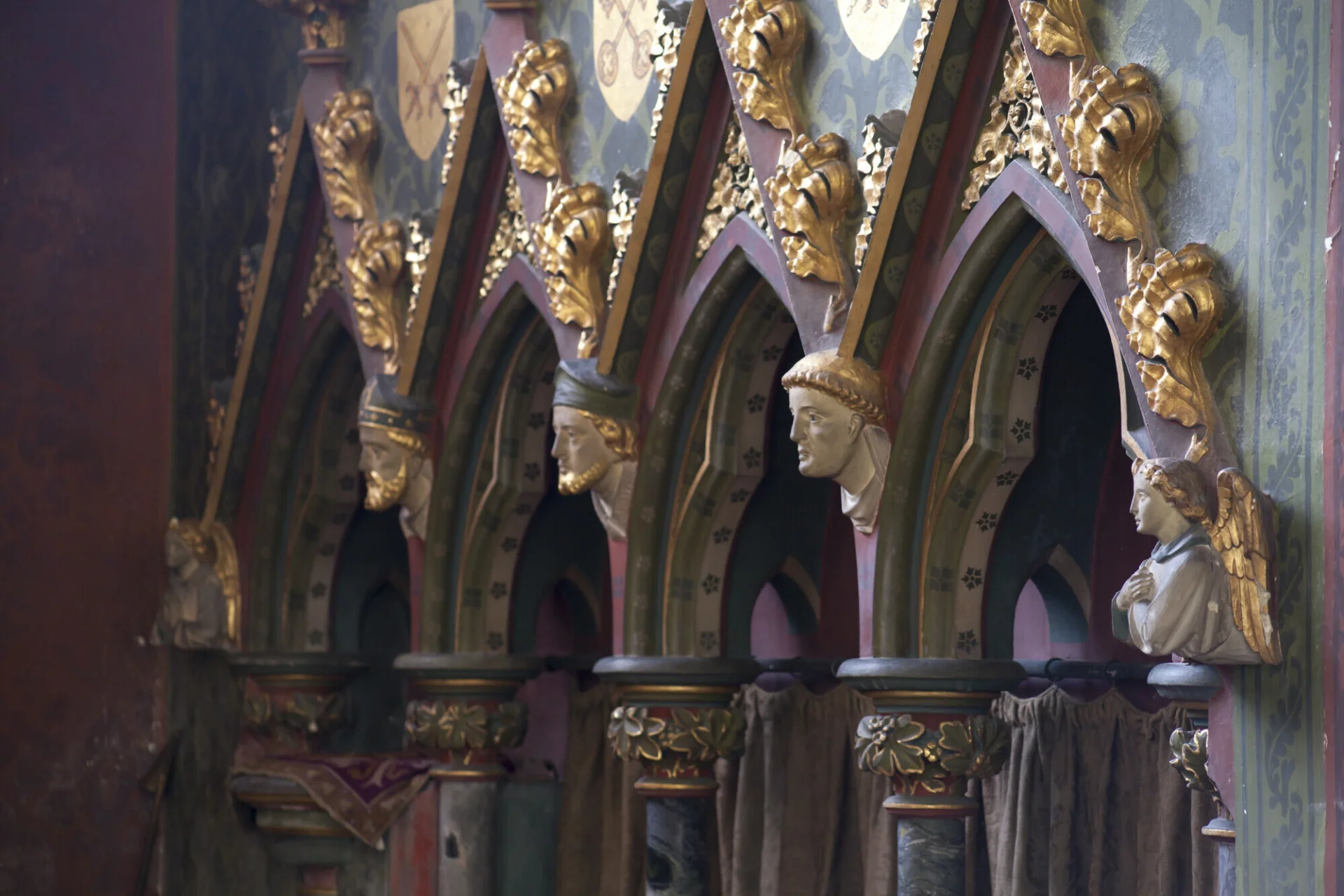 Image of richly decorated sedilia painted with red and green stencilling and decorated with tiny life-like painted corbel heads and golden flourishes.