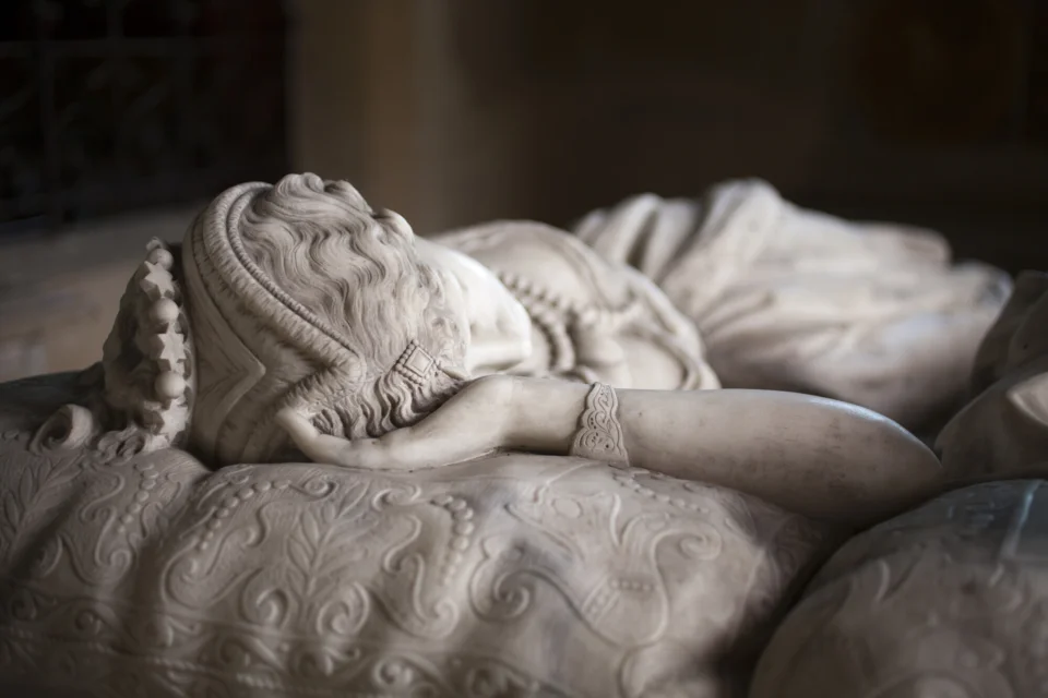 Close-up photograph of the effigy of Adeline de Horsey, depicted as a beautiful woman on her thirties, lying in her back, her face turned toward her husband, resting in her right hand. She can be seen as if we are standing at the head of her bed, with a focus on the top of her head and the elaborate pattern of her pillow. She has wavy hair and wears a richly decorated headdress including a chain of star-shaped beads.