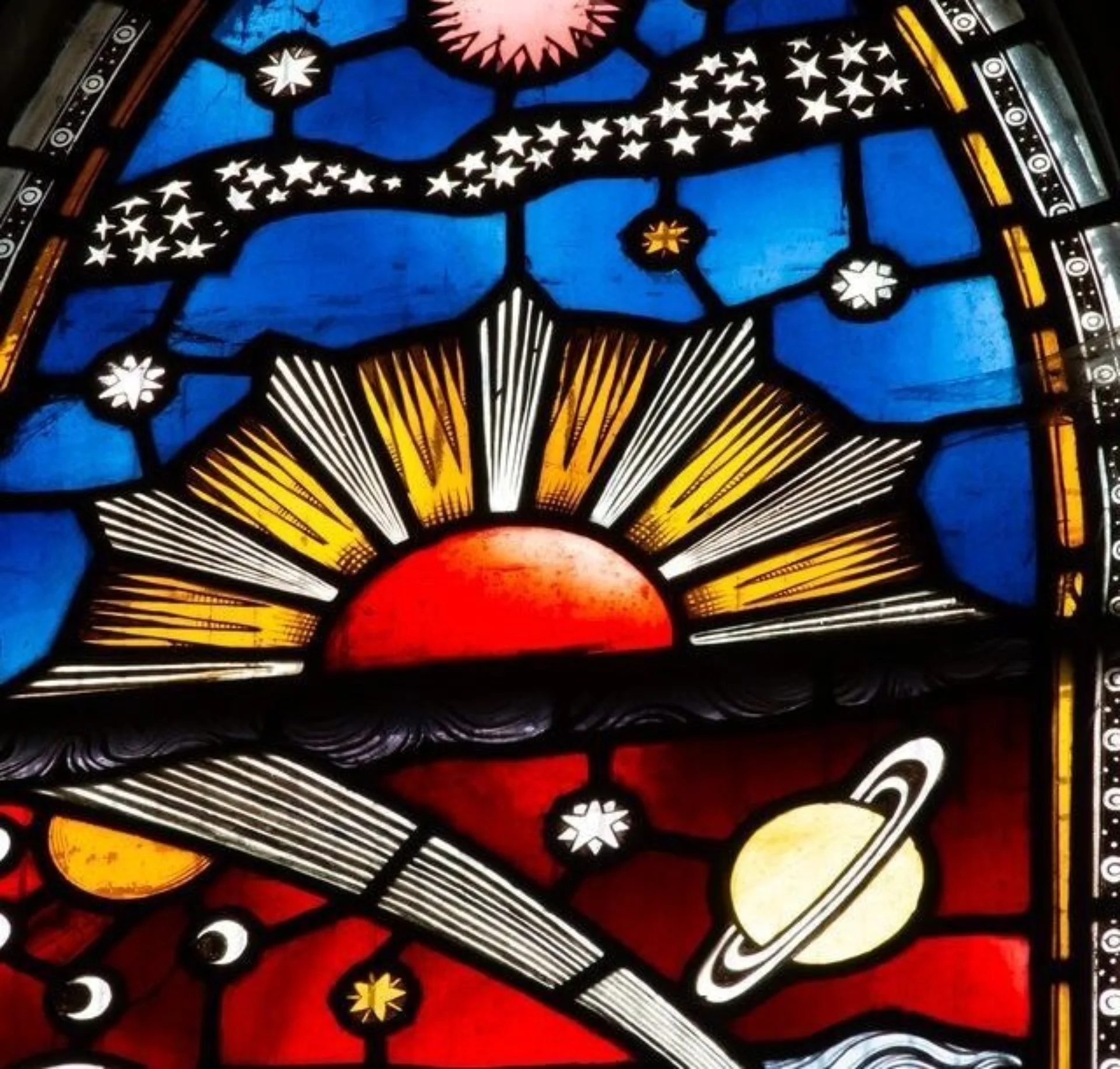 Stained glass window depicting a red sun on blue ground and stars, moon, planets and comets on red ground underneath.