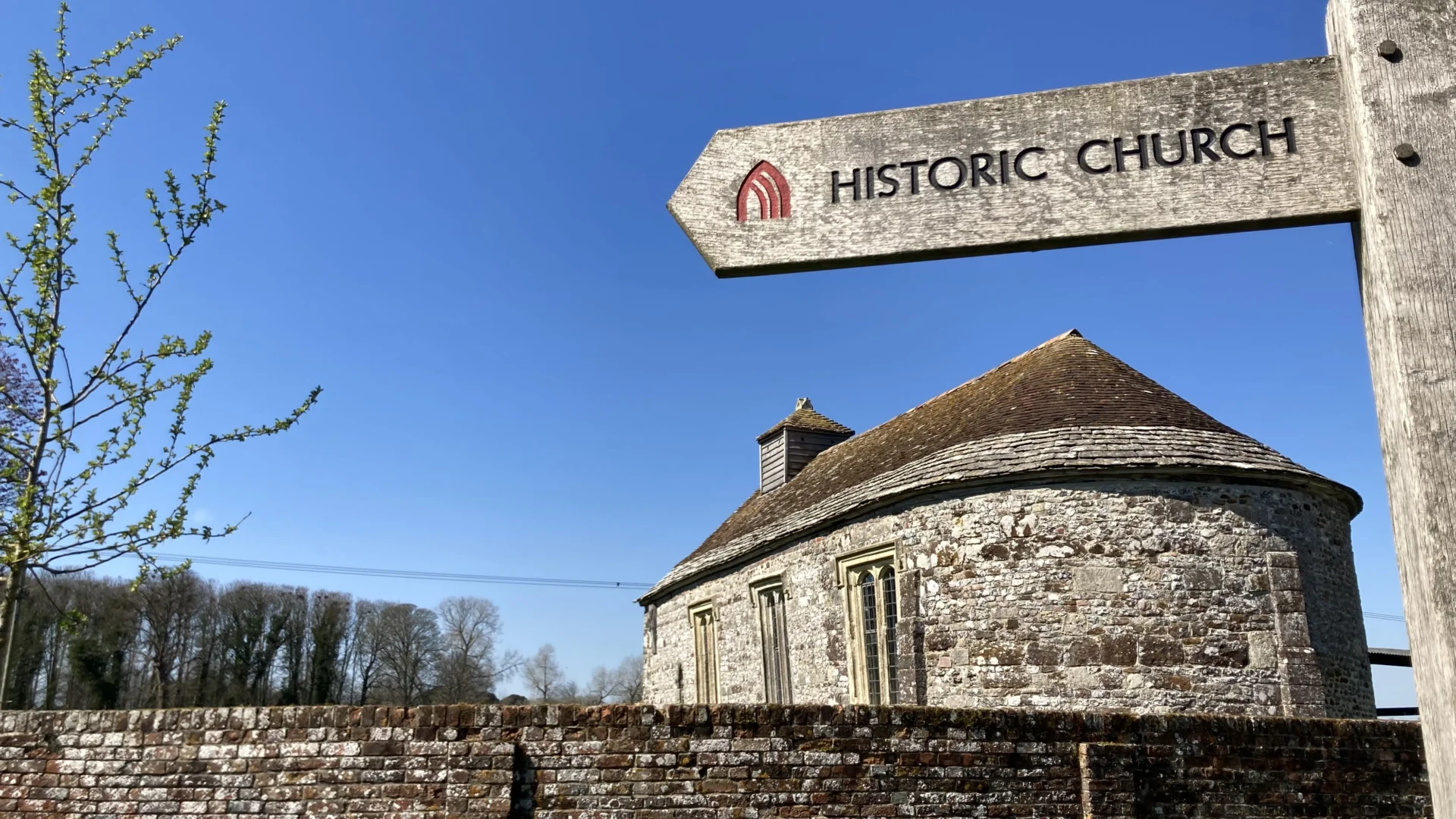 Side view of the exterior of the stone built church from behind stone wall wall with historic church sign