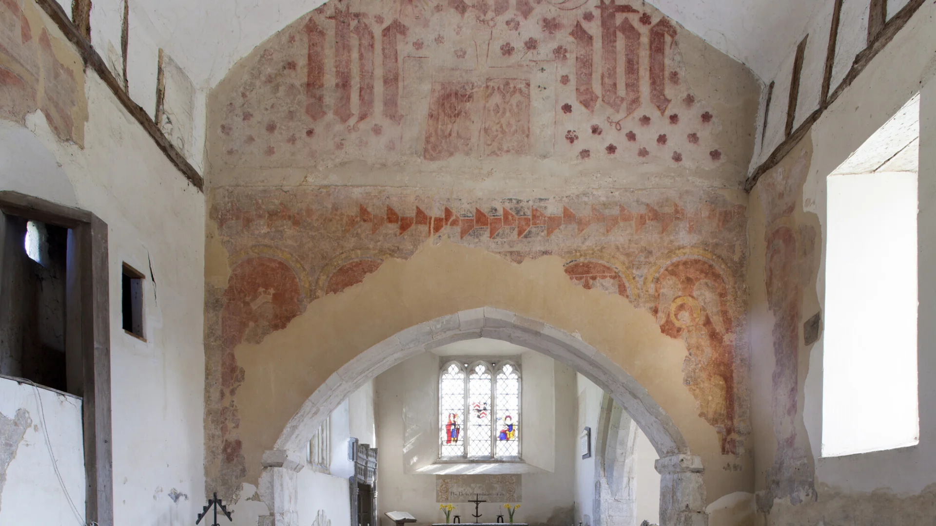 A narrow altar with two wooden pews, above sits n arch with colourful (golds and reds) wall paintings