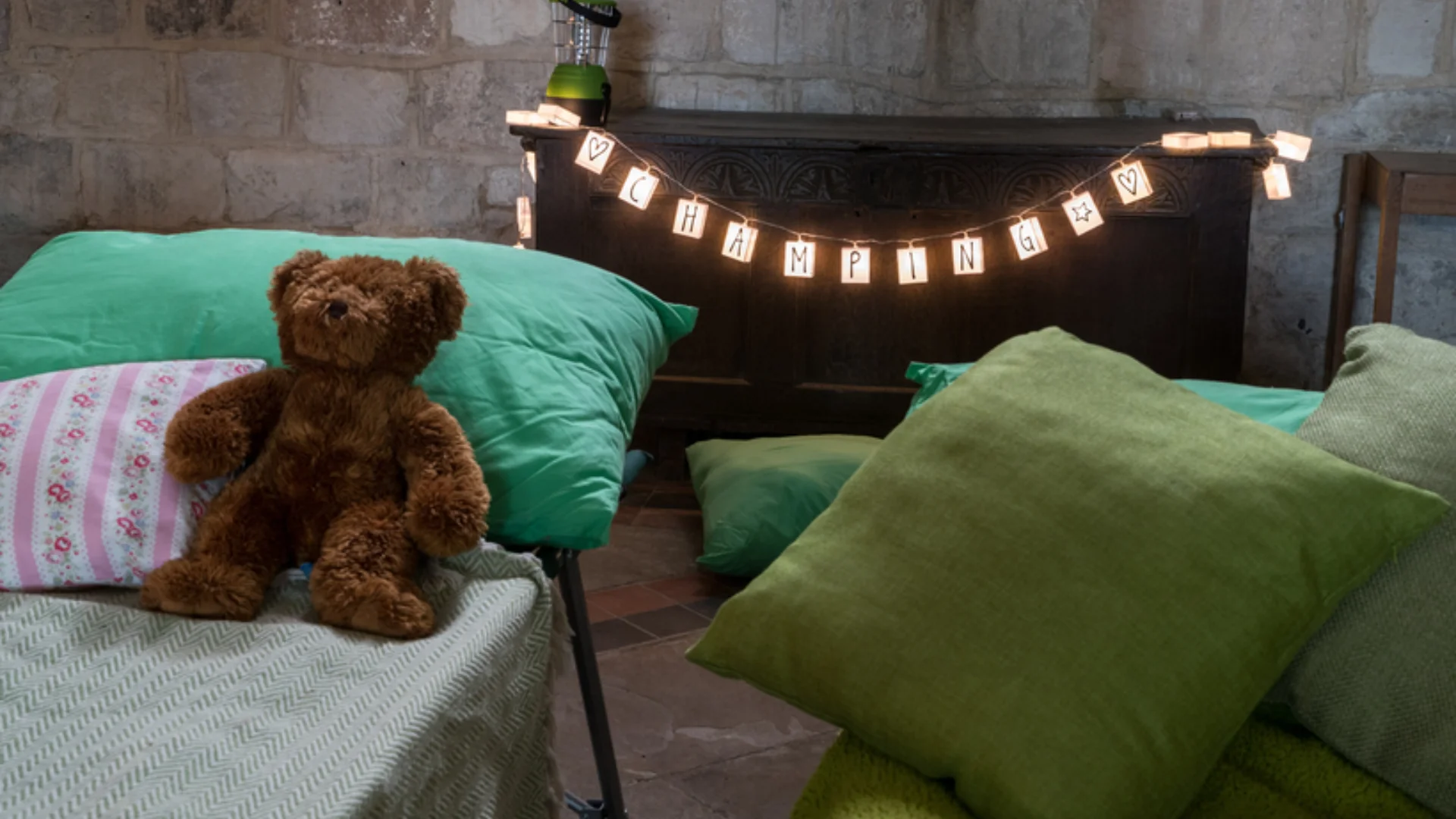 Cosy scene of camp bed with green cushion, pink and white floral cushion and warm brown teddy bear with green sheets. In background fairy lights are strewn over dark wooden table which also has a camp light on. To right, green cushions on green upholstered bench.