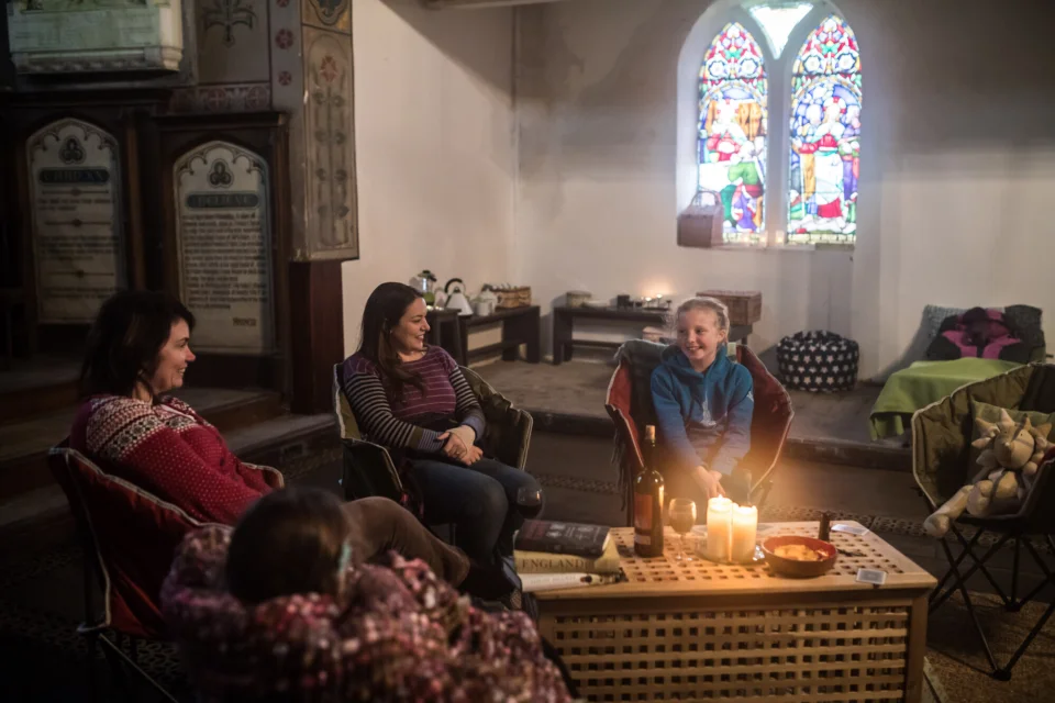 Three people sitting on chairs around a coffee table with a decorative screen to the left and stained glass window behind.