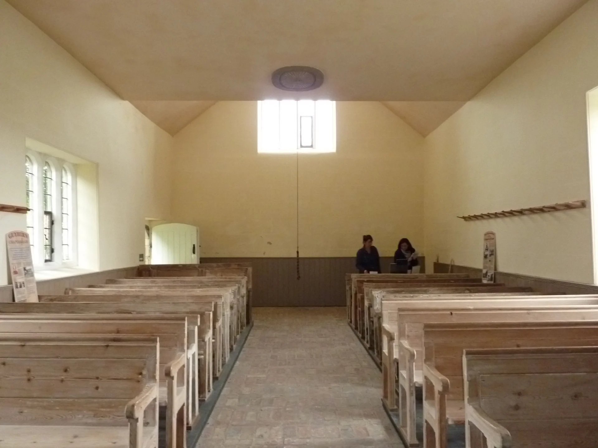 brick and stone nave with plain glass windows and original narrow pews. Two people sit at the back