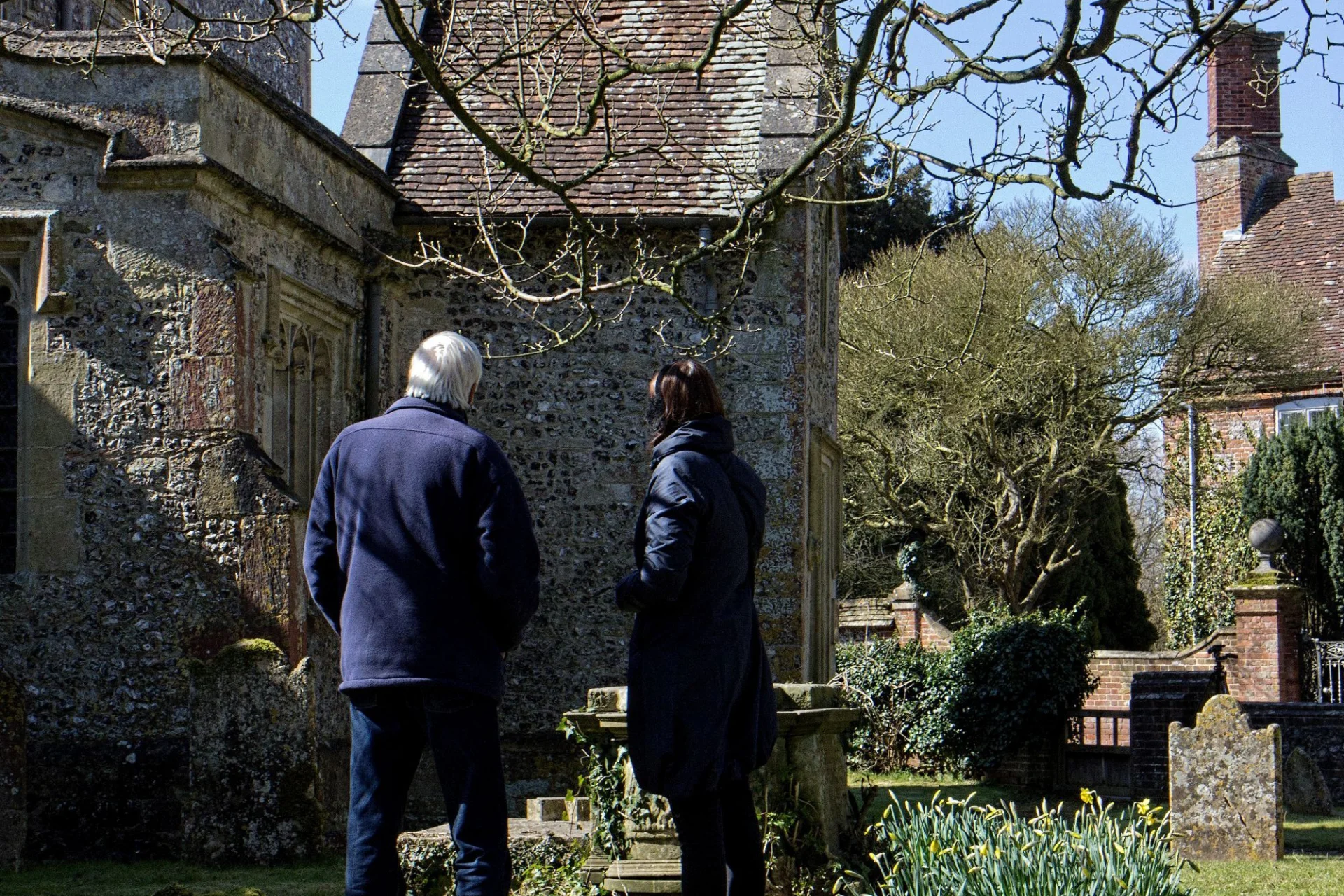 Two people in winter coats have their back to the camera as they look up at an old stone church.