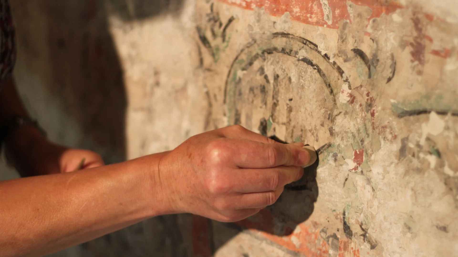 A hand shown holding a delicate paintbrush, working on the conservation of the wall art