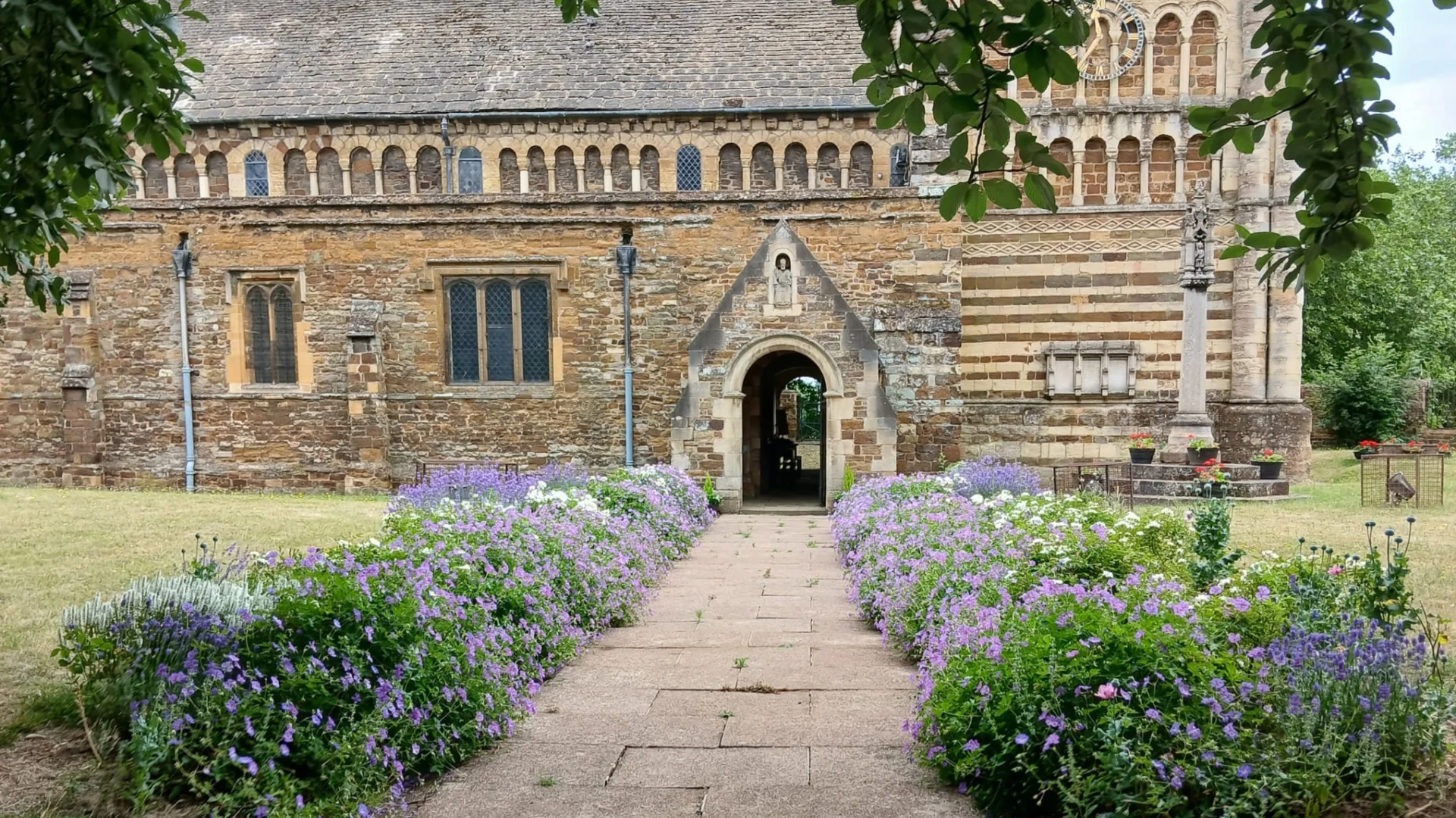 looking down the churchyard path, lined by purple flowering plants, towards the church with its door open