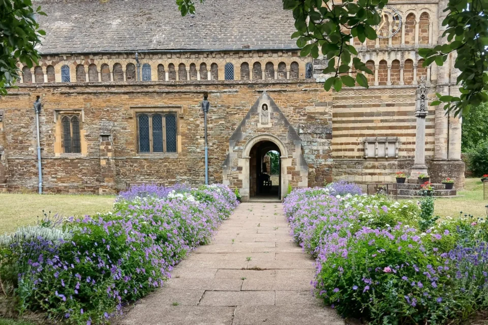 looking down the churchyard path, lined by purple flowering plants, towards the church with its door open