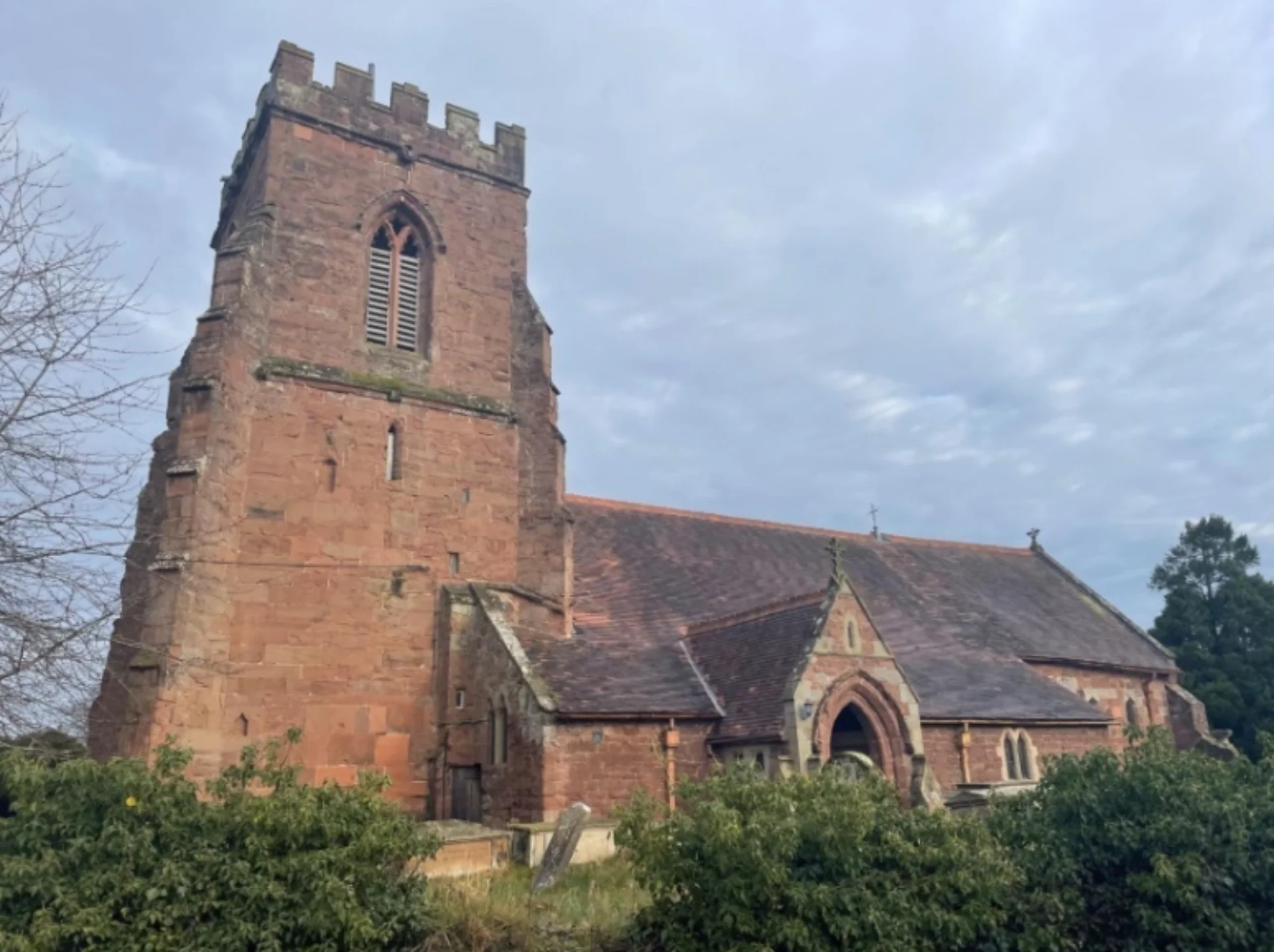 A sandstone medieval church with a square tower situated in a churchyard with greenery.
