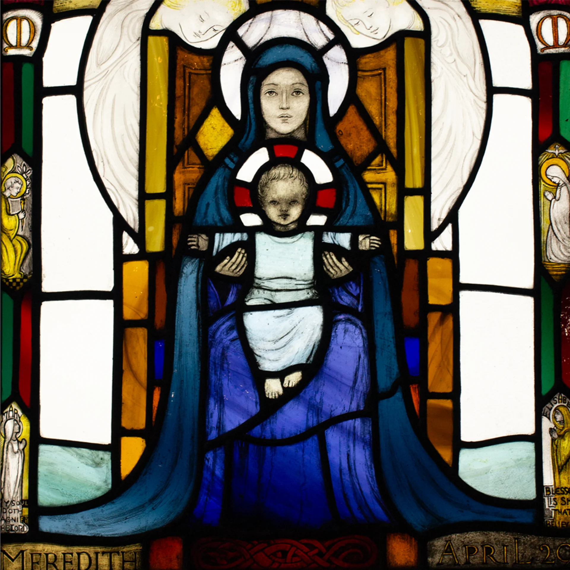 A square stained glass window panel with the virgin in blue dress holding the Christ child on her lap.