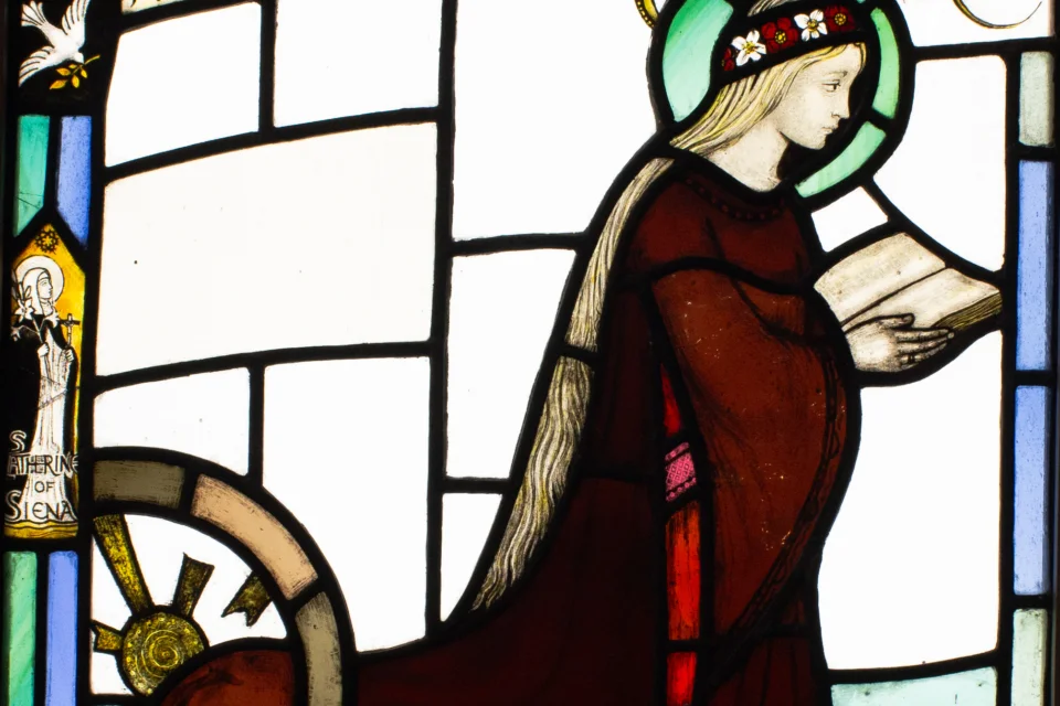Square window panel depicting St. Catharine of Alexandria as a young woman in red dress, flower crown, kneeling, reading