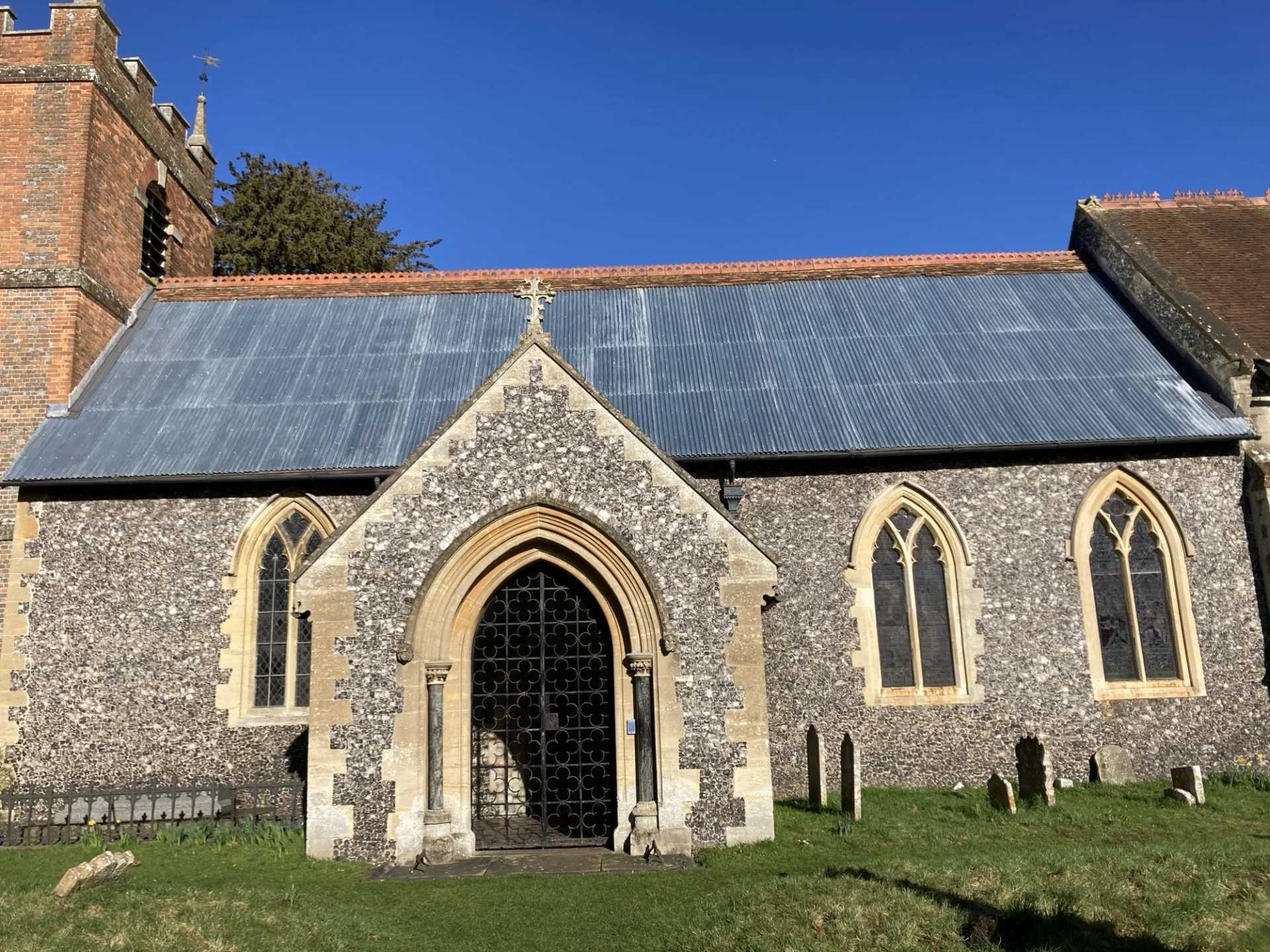 Full view of St. Bartholomew's, Lower Basildon, a historich church with flint covered porch and nave, three tall nave windows, a crenellated red brick tower and a corrugated iron roof.