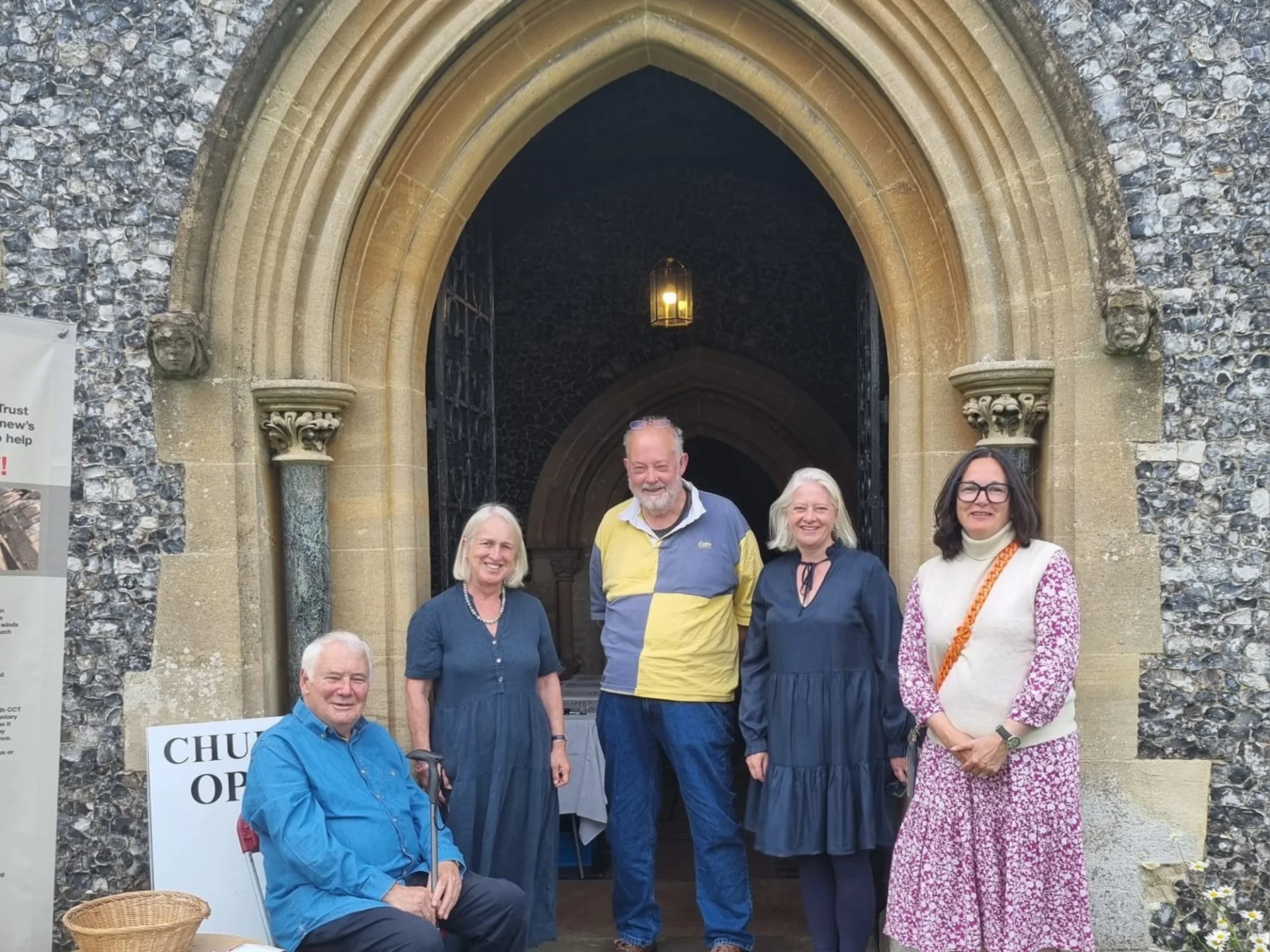 Five people posing for a photograph in front of a pointed church archway. All are smiling, a man on the left in blue shirt is sitting down, to his right stands a woman in blue dress, next to her a tall man in jeans and blue and yellow checkered jumper, to his right another woman in a blue dress almost identical to the first woman's dress and to her right stands a woman in a pink dress, with a white vest top. All have grey hair, except the woman on the far right, who has black hair.