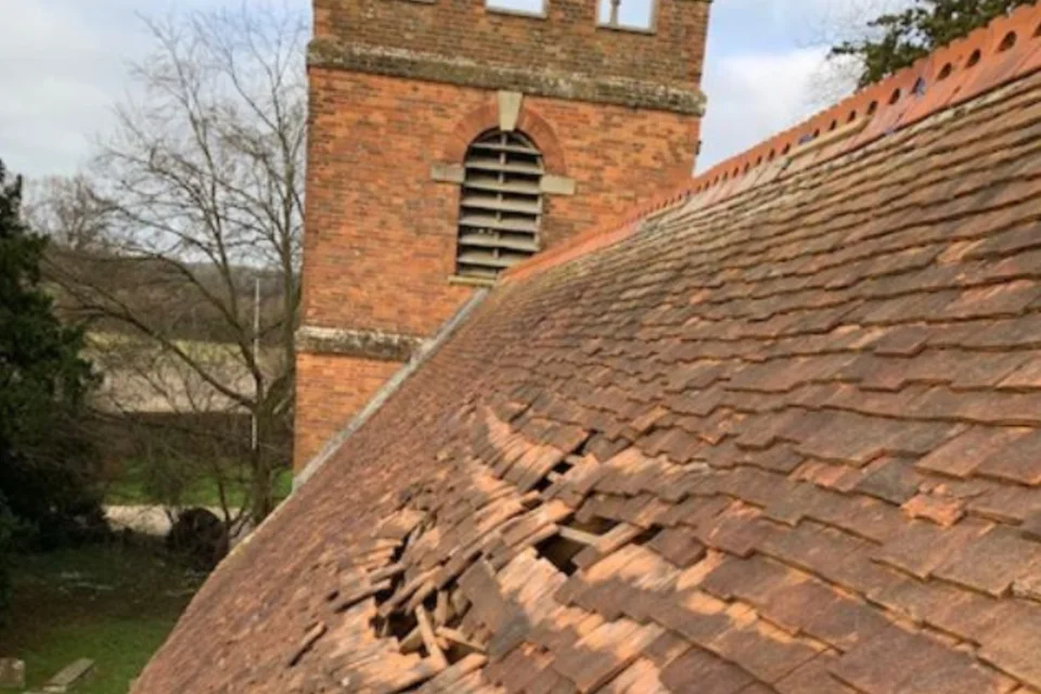 Close-up view of a red tile church roof in poor condition with several broken and slipped tiles opening up holes in the roof. A crenellated red brick church tower can be seen in the background.