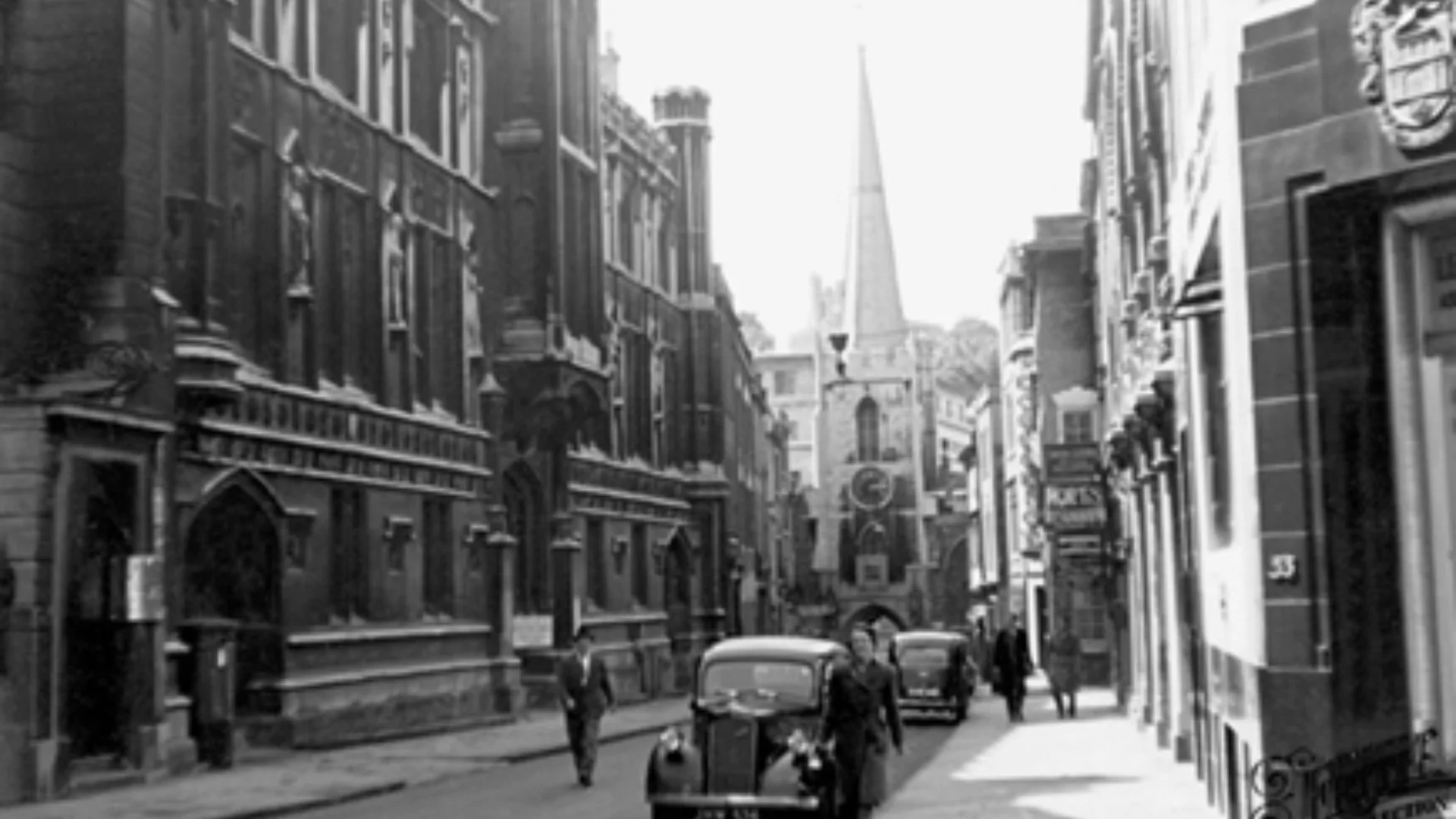 View of Broad Street in Bristol with hotels and shops on either side of the street and vintage cars parked at the side of the road. The tower and spire of Bristol St John can be seen in the background.