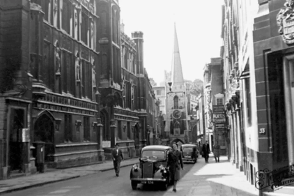 View of Broad Street in Bristol with hotels and shops on either side of the street and vintage cars parked at the side of the road. The tower and spire of Bristol St John can be seen in the background.