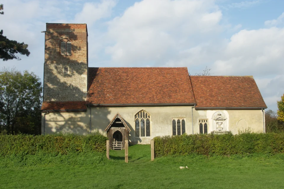 Image of church exterior flint-and-brick with red tiled roof in an field surrounded by a thick hedge.