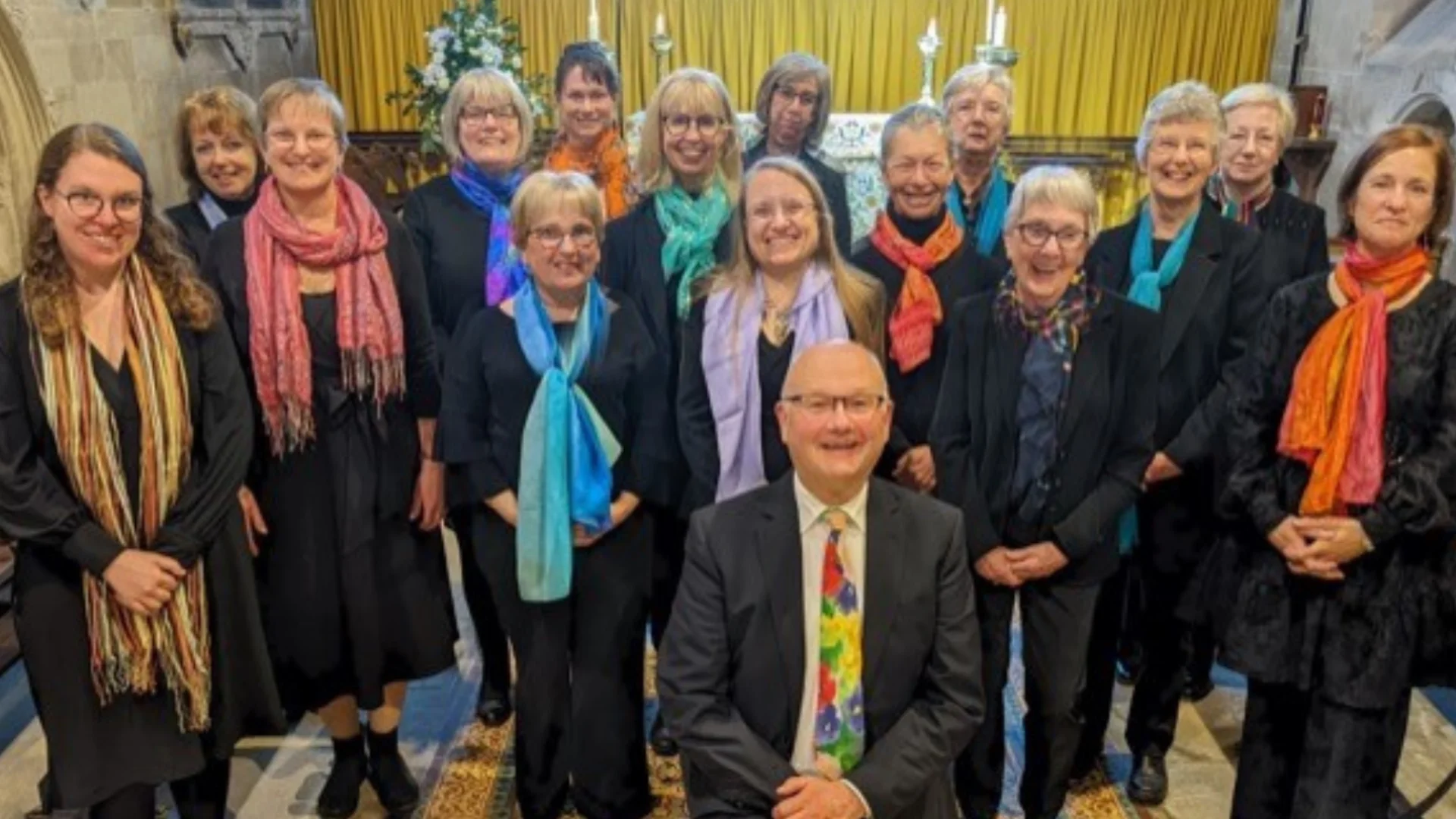 Group of 16 singers pose for a photograph, they are stood up except for a male member of the choir kneeling at the front.
