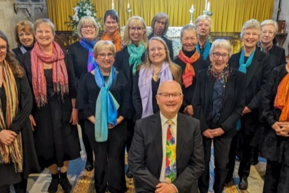 Group of 16 singers pose for a photograph, they are stood up except for a male member of the choir kneeling at the front.