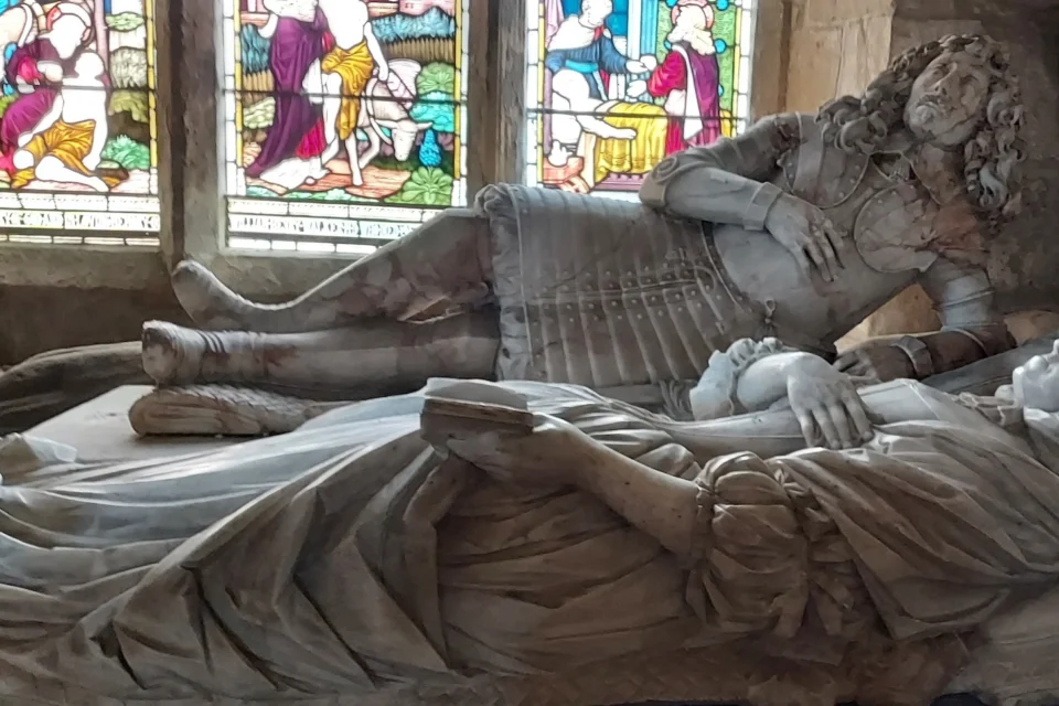 Man and woman captured in stone, man in full armour lays on side propped up on one arm, with the other to his chest. Woman lies next to him in robes, holding book in one hand and with the other to their chest.