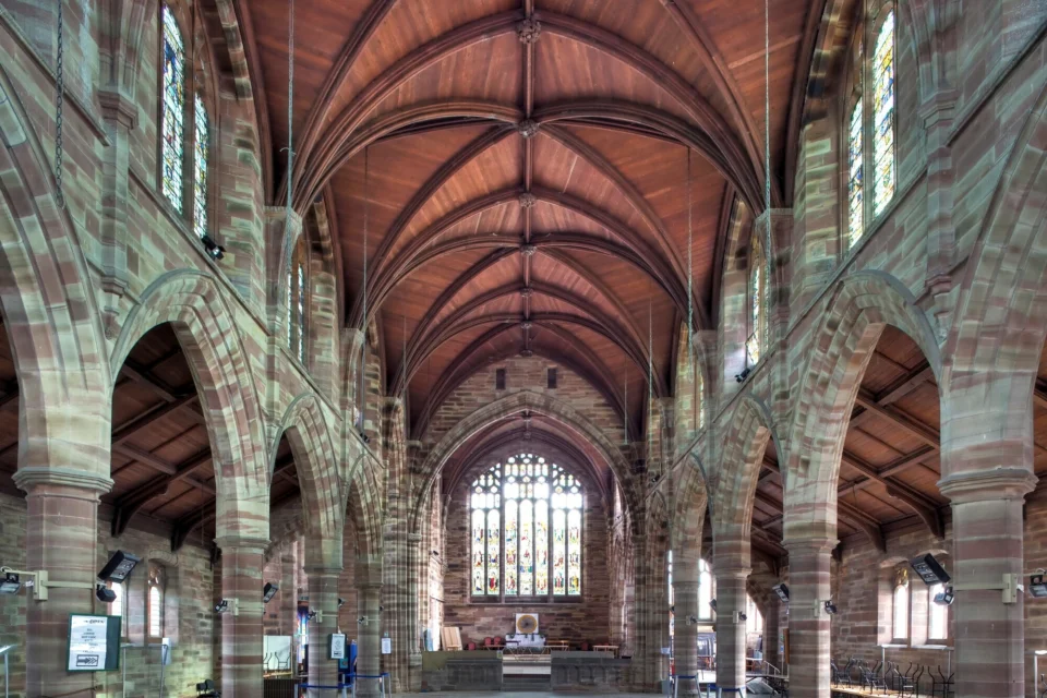A stunning stone Nave with arches on each side and a timber roof. A stained glass window sits above the alter.