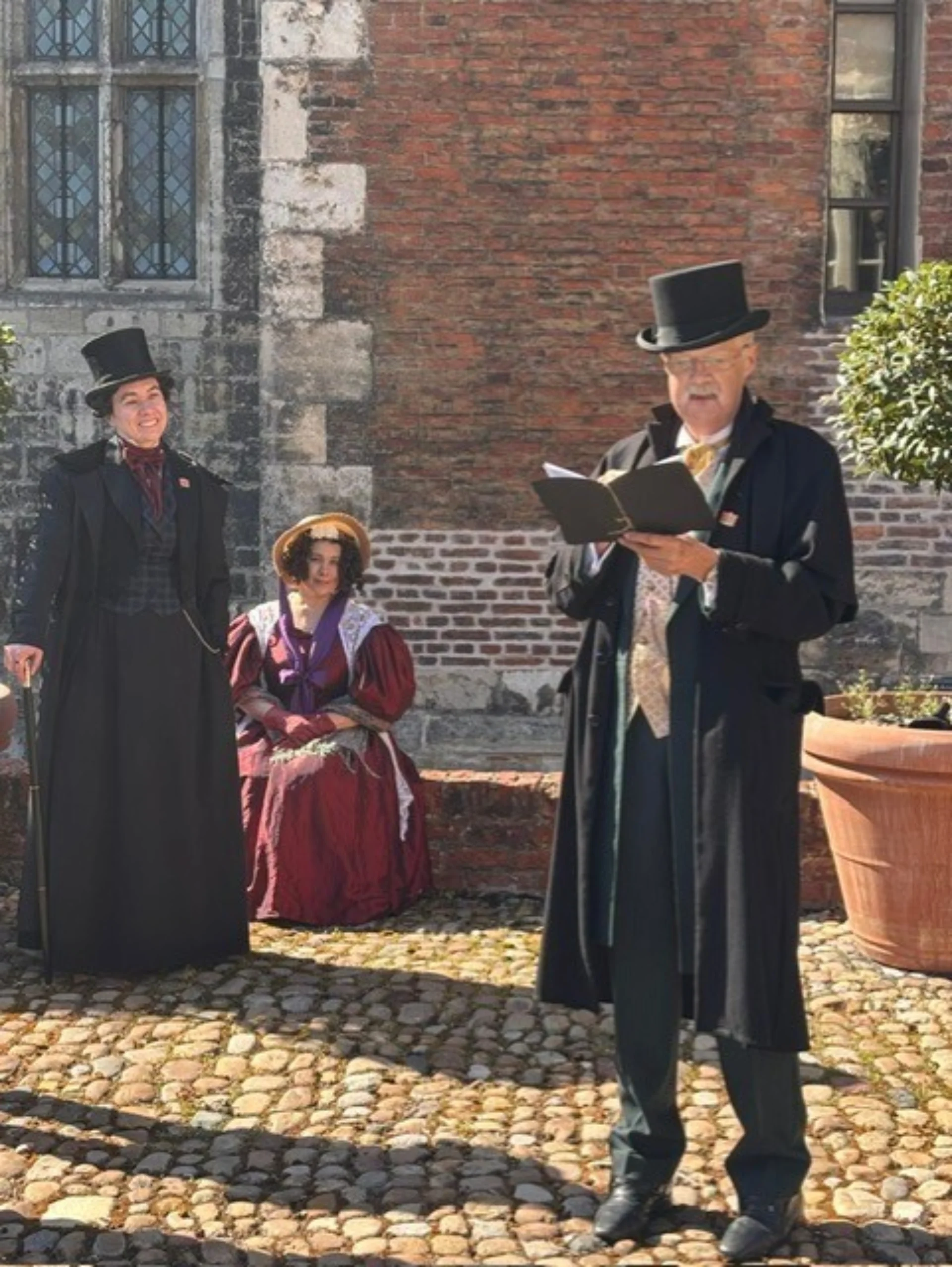 Two women dressed as Anne Lister and Ann Walker listening to a man in a top hat read from a book.