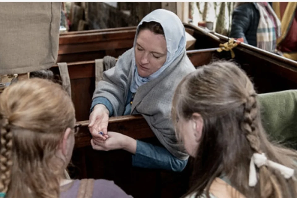 Woman wearing Viking style blue dress and white headscarf talking to two girls with their backs to the camera