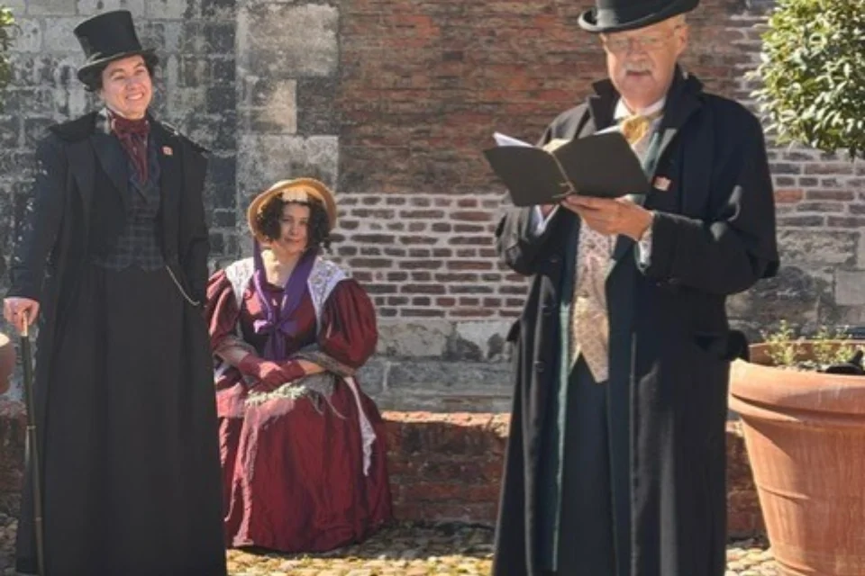 Two women dressed as Anne Lister and Ann Walker listening to a man in a top hat read from a book.