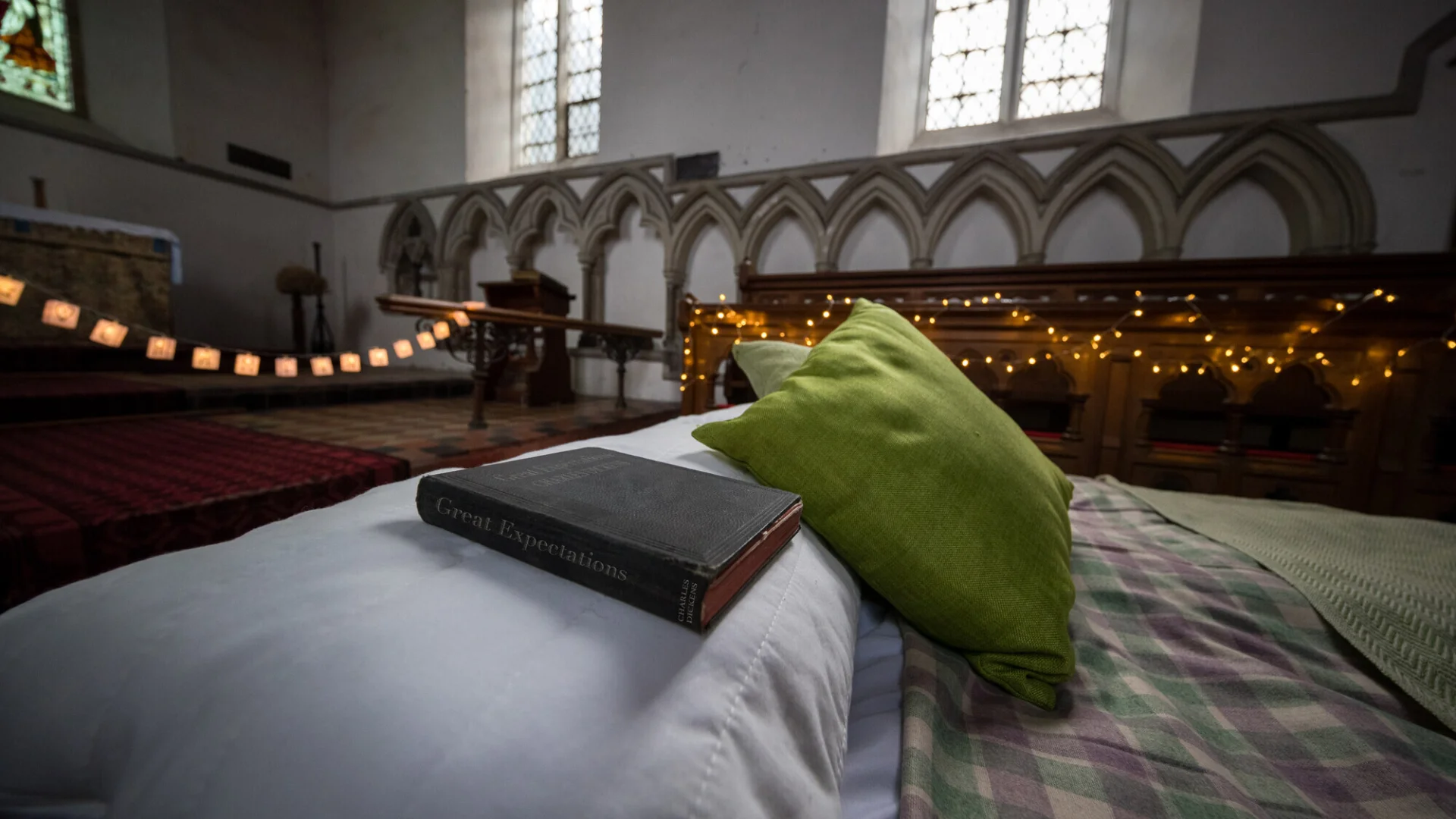 Camp bed with blanket, pillow, green cushion and antique copy of Great Expectations, in background church windows with fairy lights