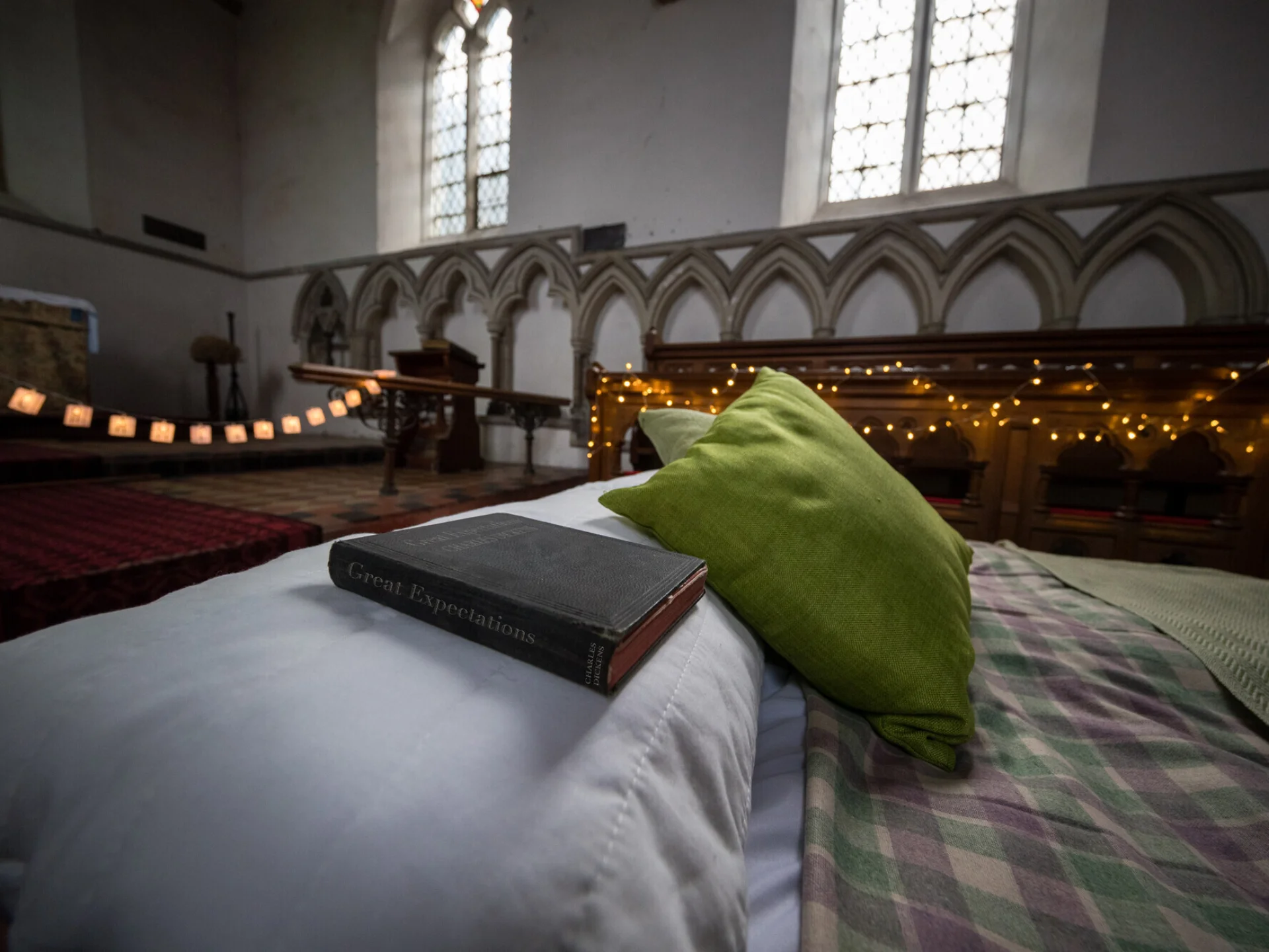 Camp bed with blanket, pillow, green cushion and antique copy of Great Expectations, in background church windows with fairy lights