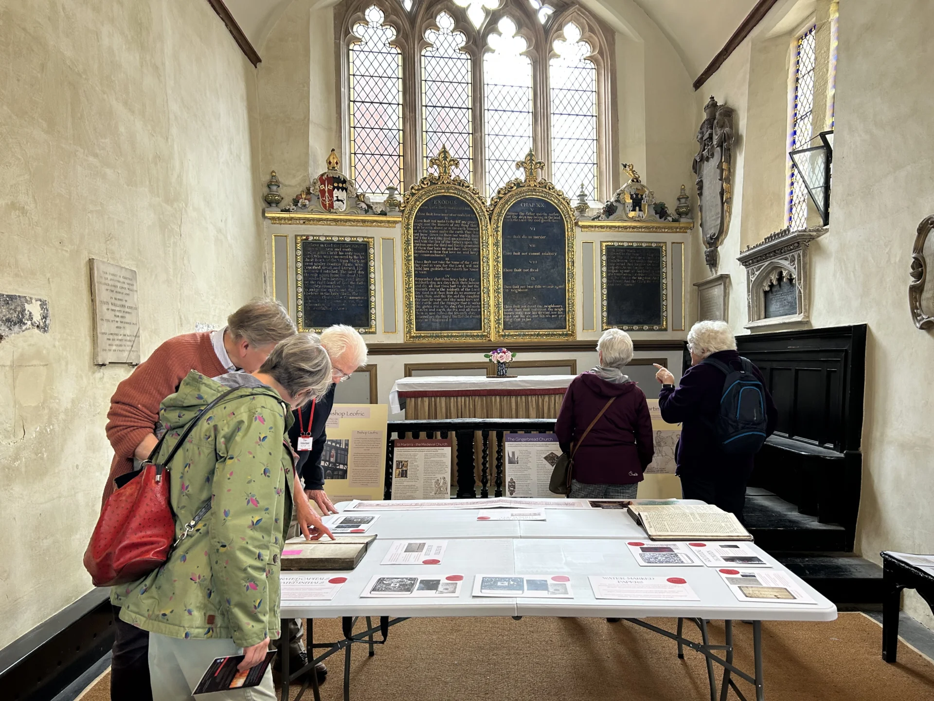 several people hover over a table with several books open. A stained glass window is to the rear.