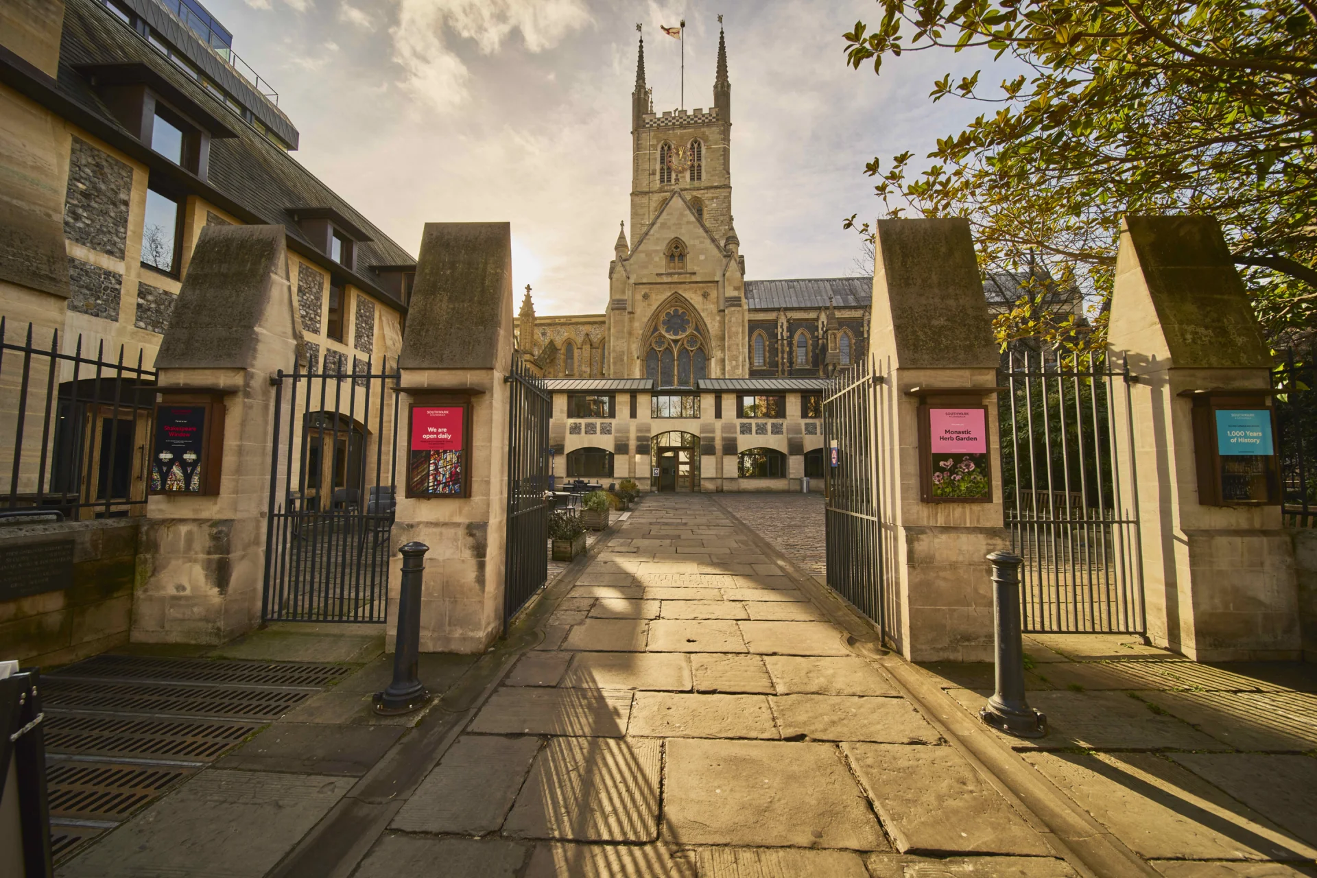 large stone slabs lead you through a iron gated entrance, with a large stone columns lined with brightly coloured posters. A cathedral standing across a courtyard dominates the sky. More modern, shorter buildings sit to the front and sides.