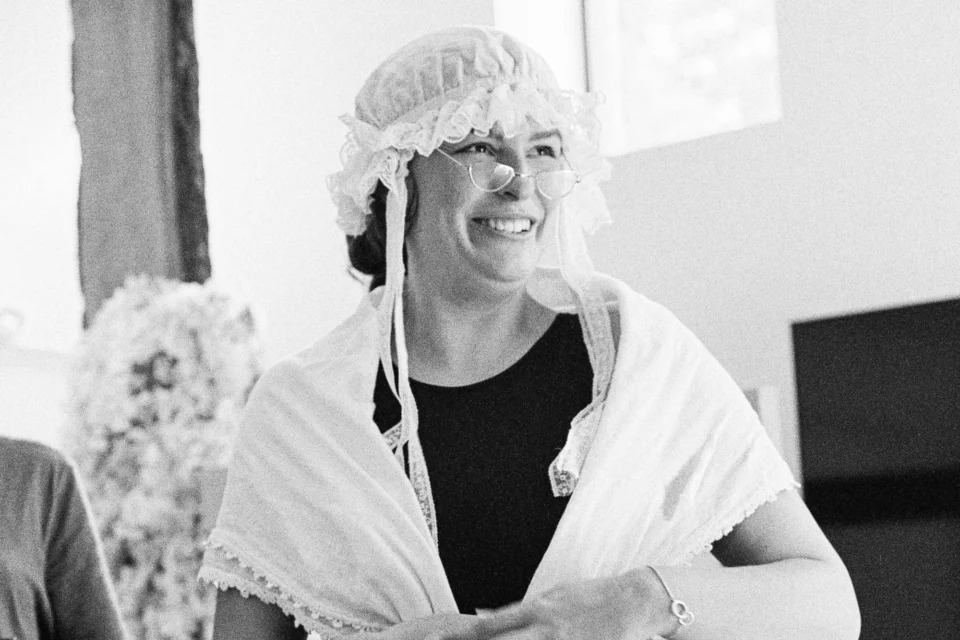 A woman in a white shawl, white lace bonnet and glasses is laughing, her hands folded in front of her stomach. Black and white photograph of theatre rehearsals.